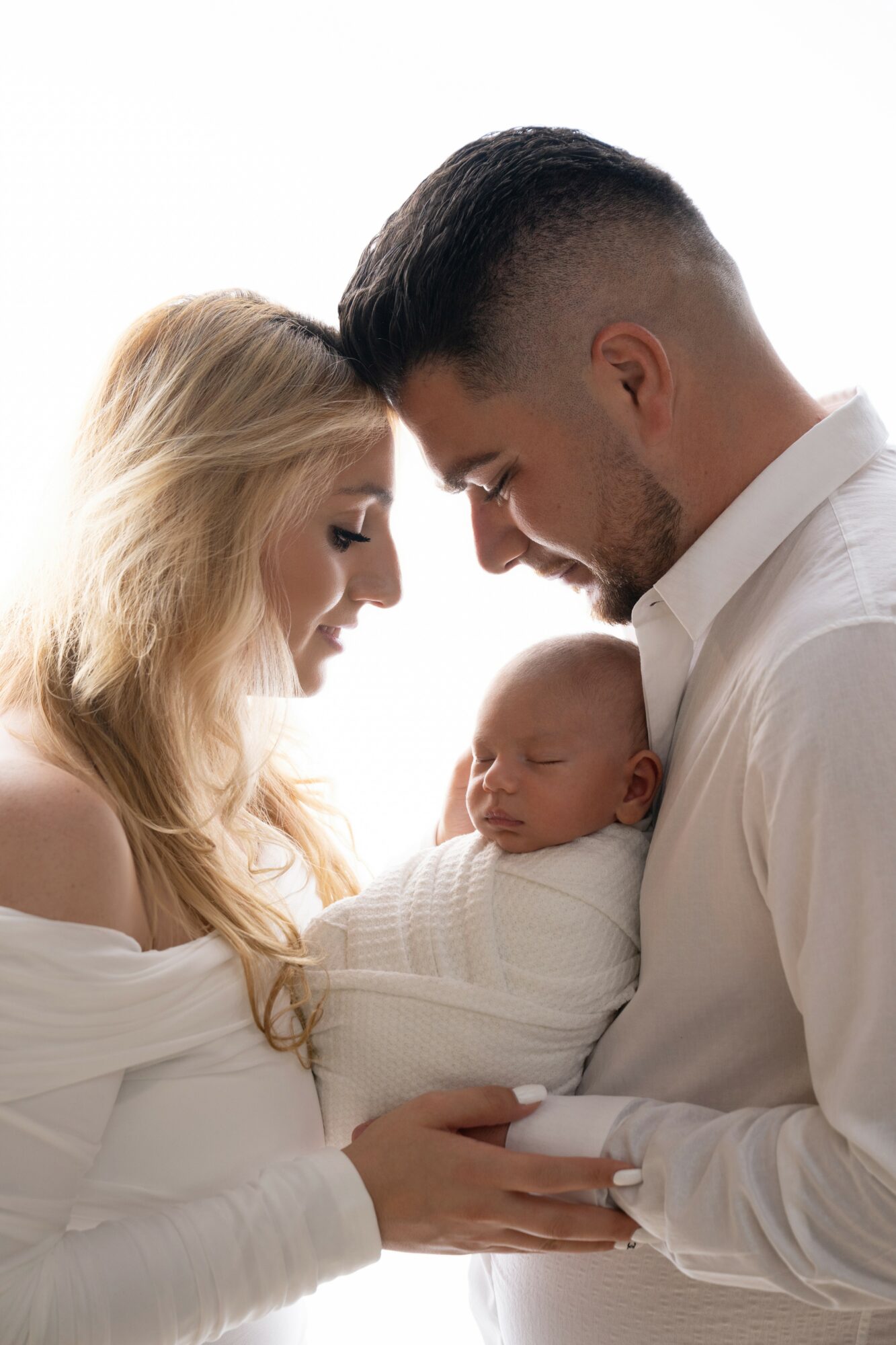 Family with a woman, man, and baby close together, smiling and touching foreheads, in a bright setting.