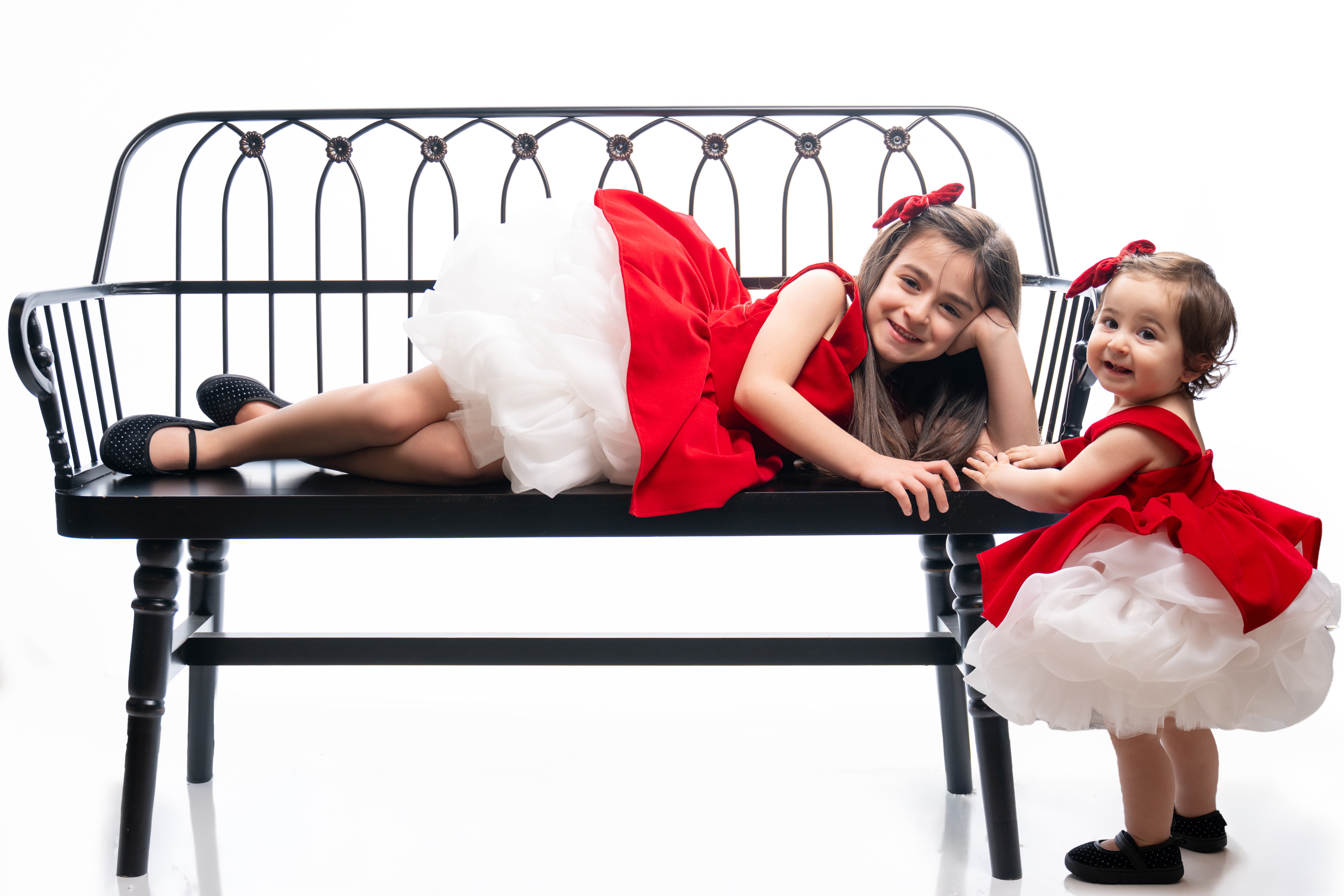 Two young girls in red dresses with white skirts lying and standing on a black bench, smiling and holding hands.