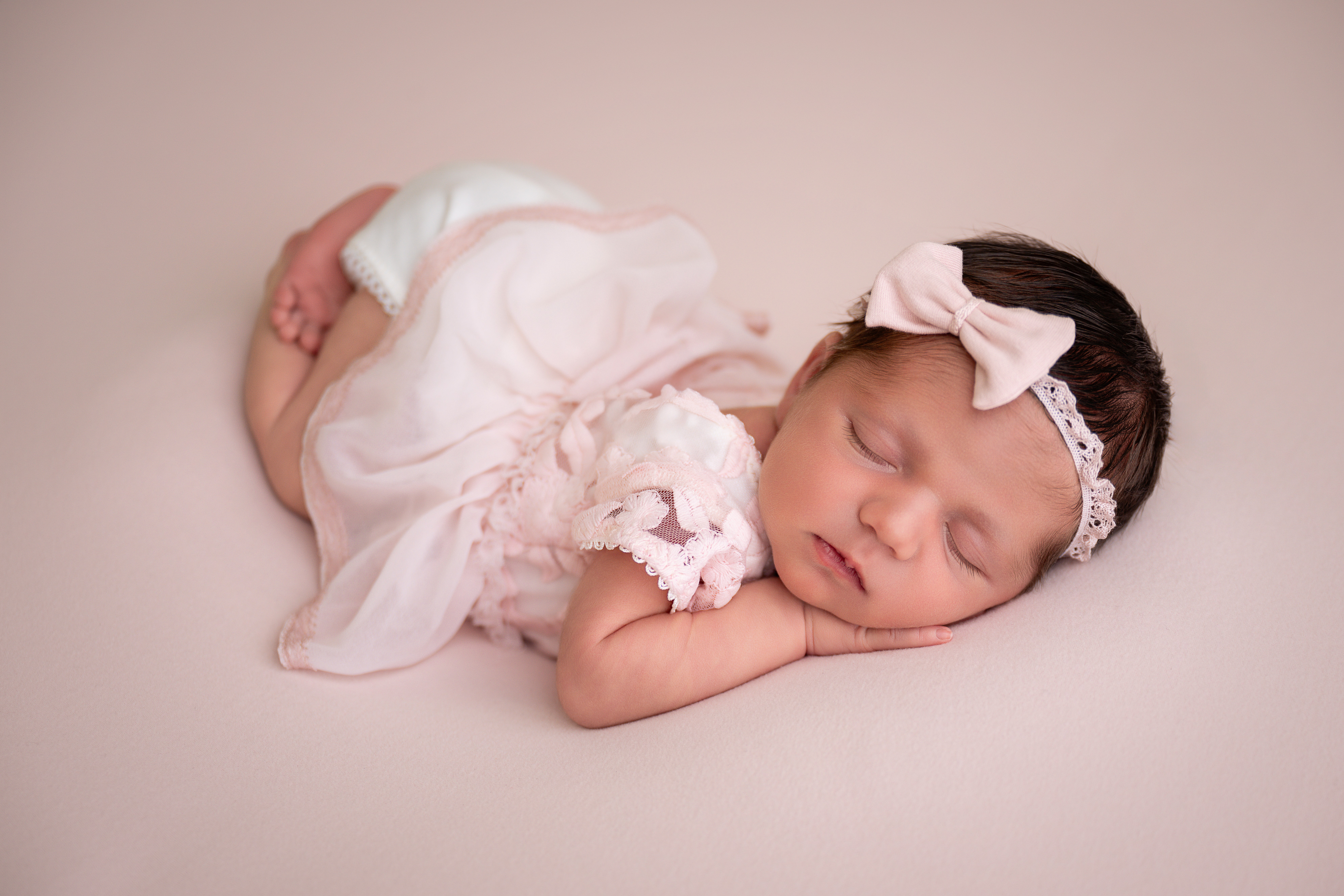 Baby girl lying on her side with eyes closed, wearing a headband and a pink dress, on a light background.