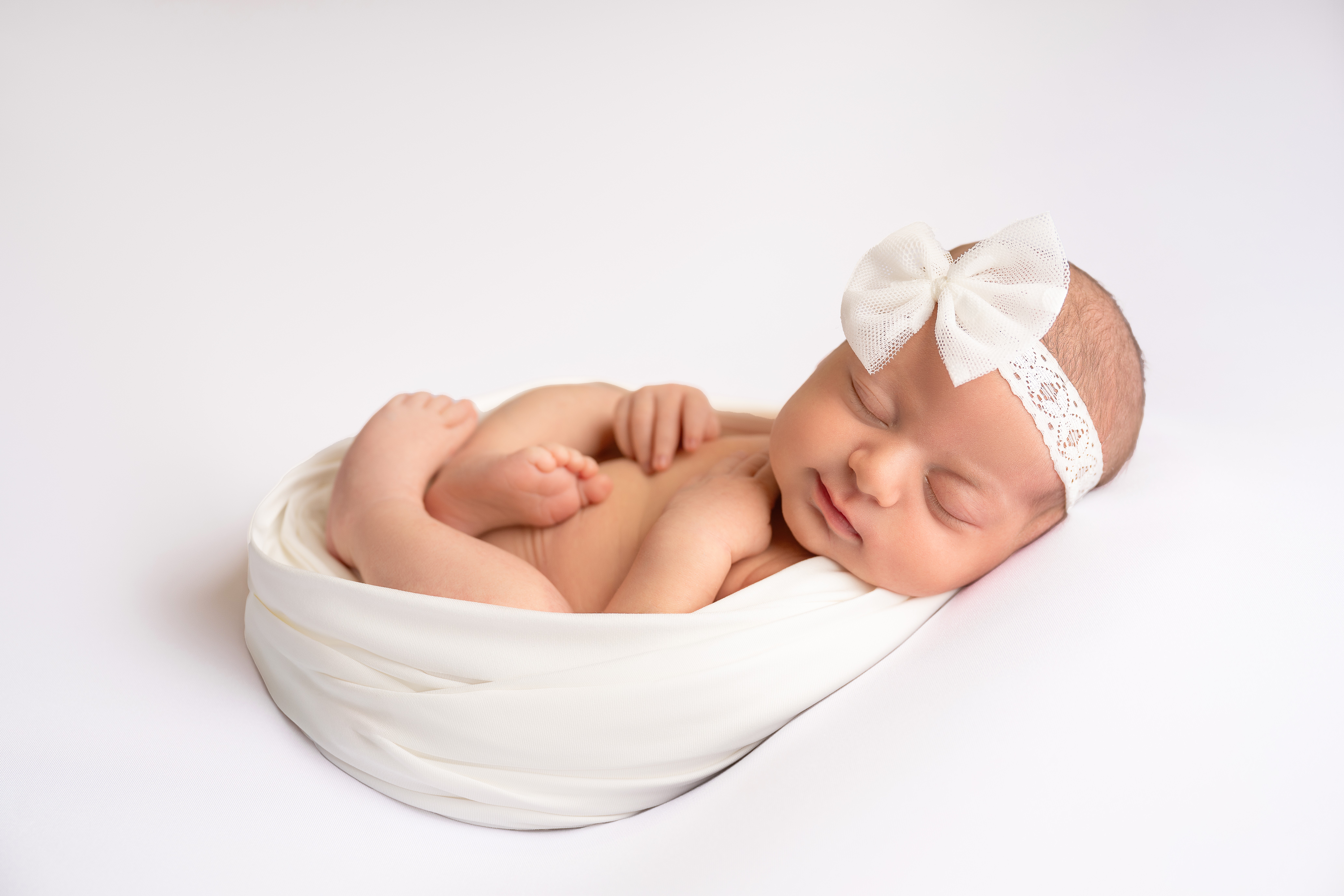 Sleeping newborn baby with a headband and bow, wrapped in white cloth, lying on a white surface.