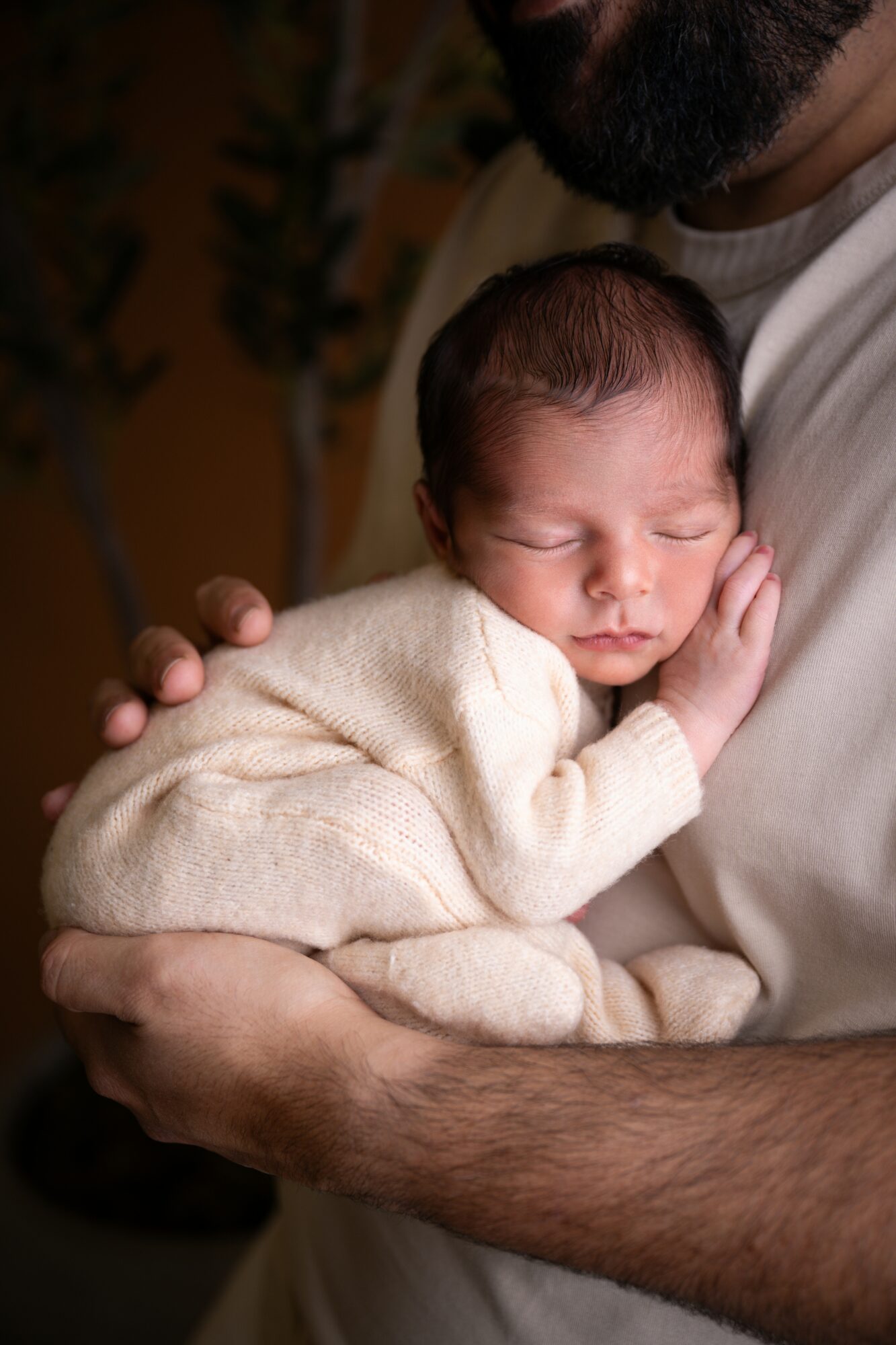 Baby with closed eyes resting on an adult's chest, held gently in arms, wearing a cream-colored sweater.