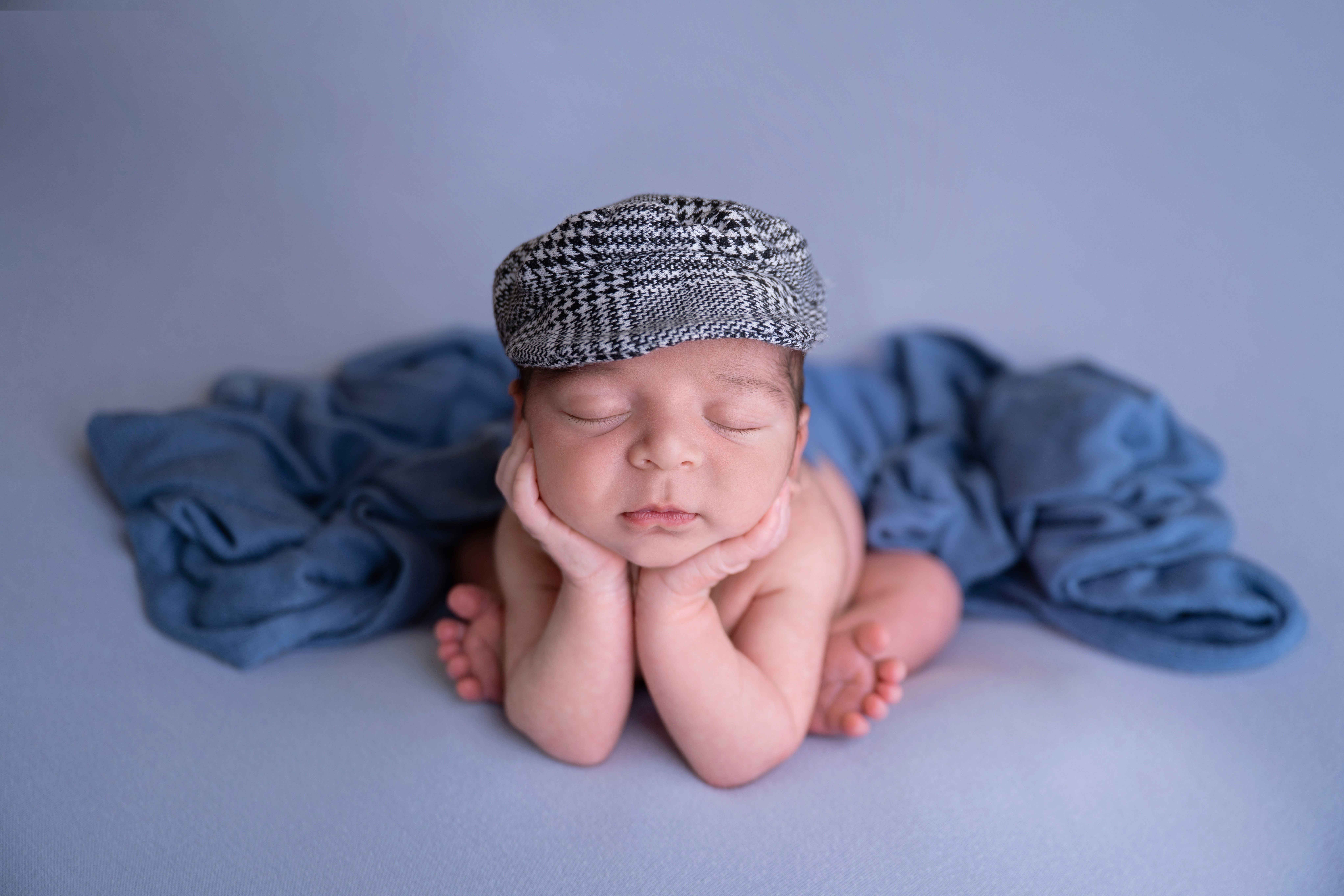 Baby with a checkered hat resting head on hands, lying on a blue blanket, eyes closed.