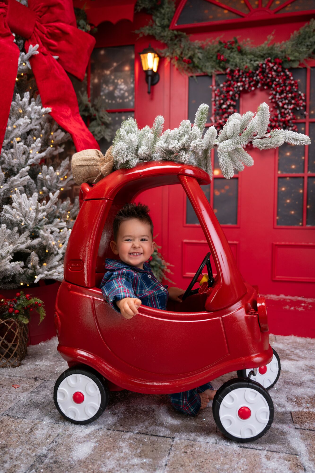Child sitting in a red toy car with a Christmas tree and festive decorations in the background.