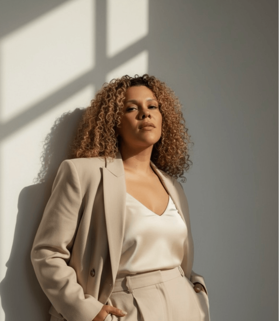 Woman with curly hair in a beige blazer and satin top standing against a wall with sunlight shadows.