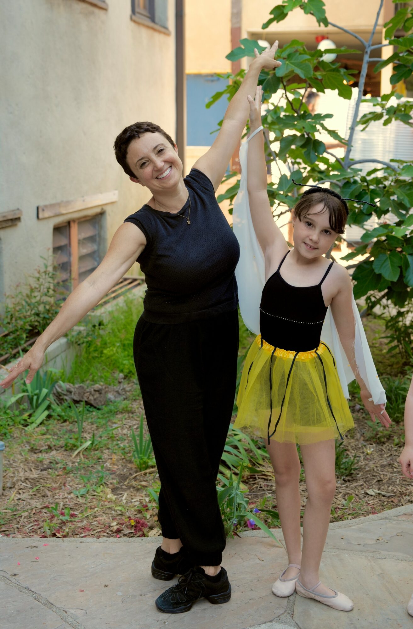Woman and girl standing outdoors near a tree, smiling, woman raising arm, girl in yellow skirt and black top, sunny day.