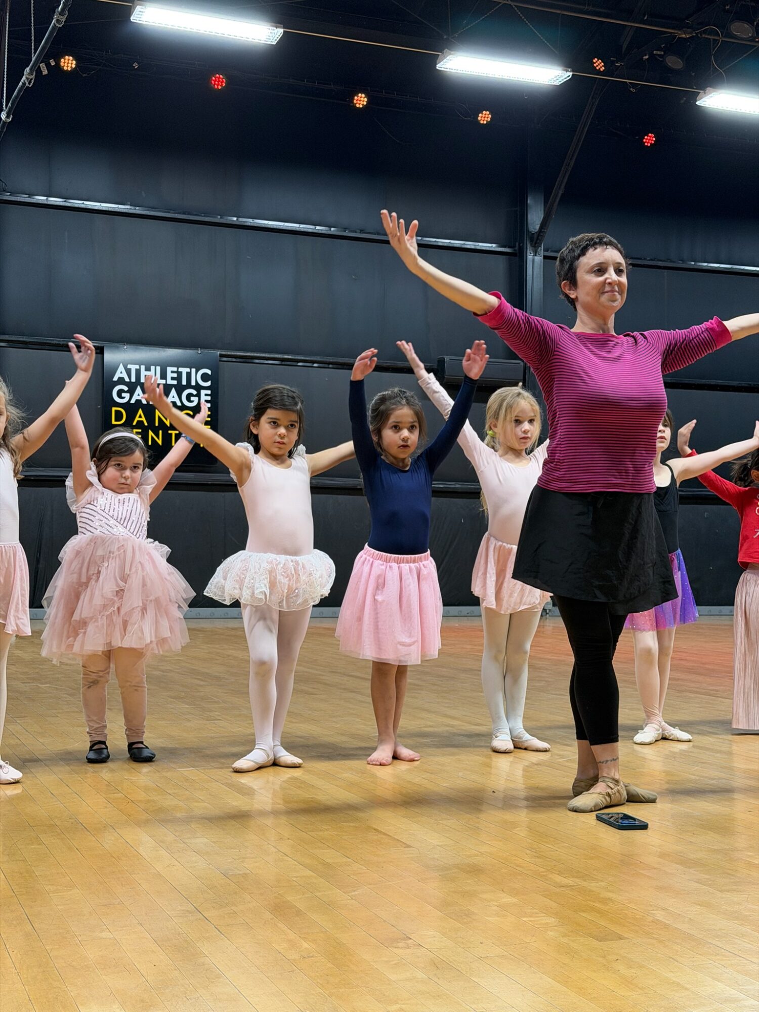 Dance instructor leading young girls in a dance class, arms raised, in a studio with wooden floor and black walls.