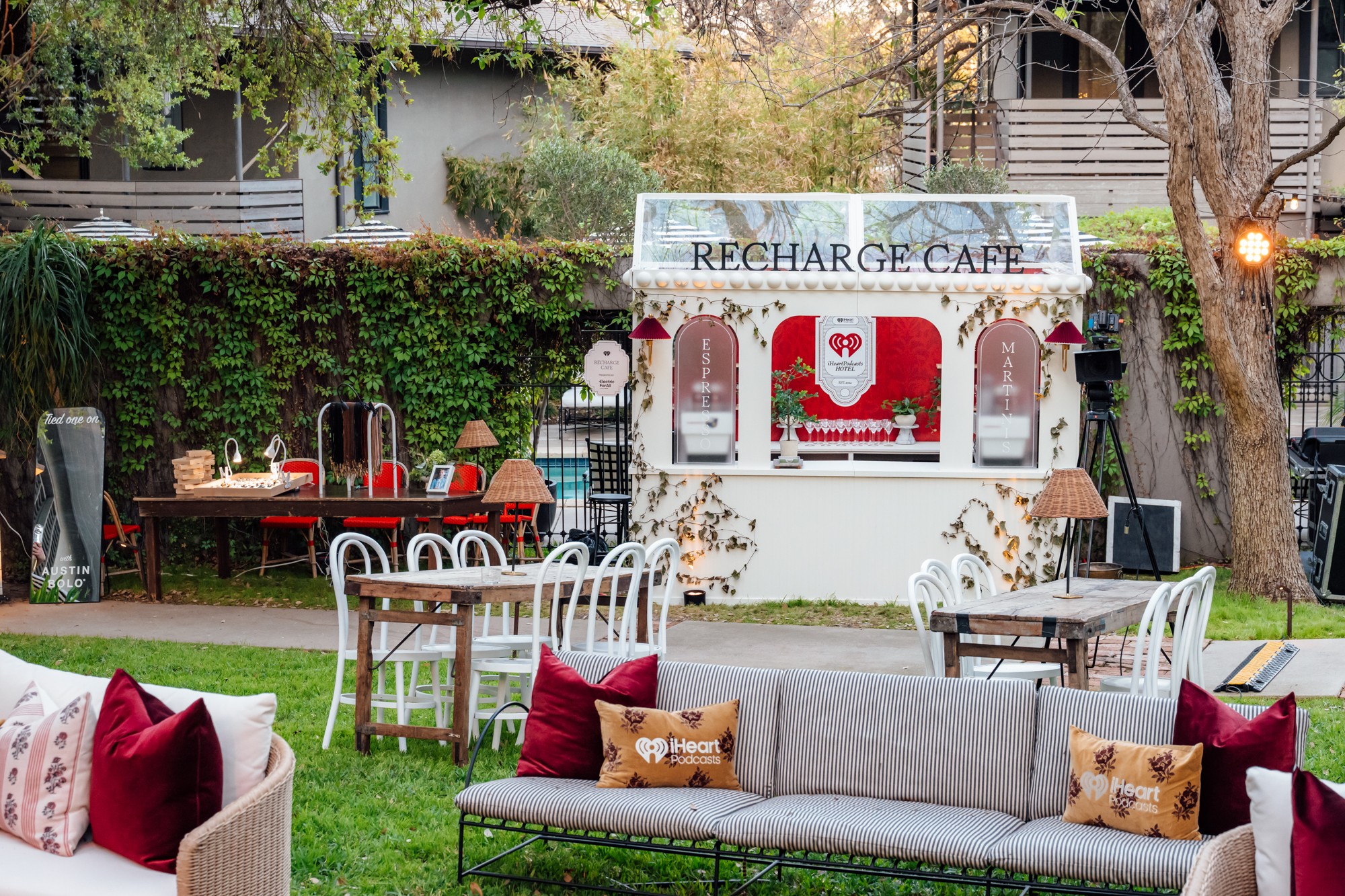 Outdoor cafe with tables, chairs, and a small food stand, surrounded by trees and greenery.