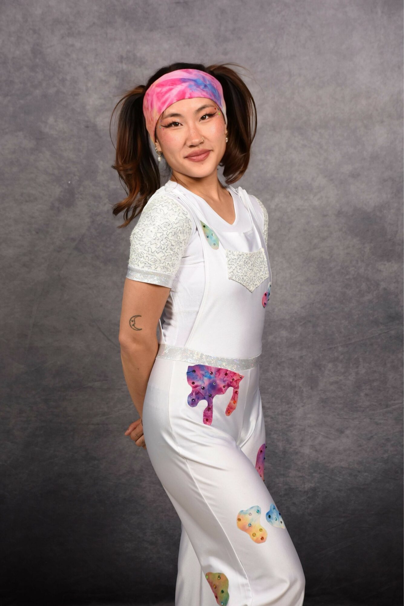 Young woman with a colorful headscarf and white overalls with colorful patches, standing against a gray background.