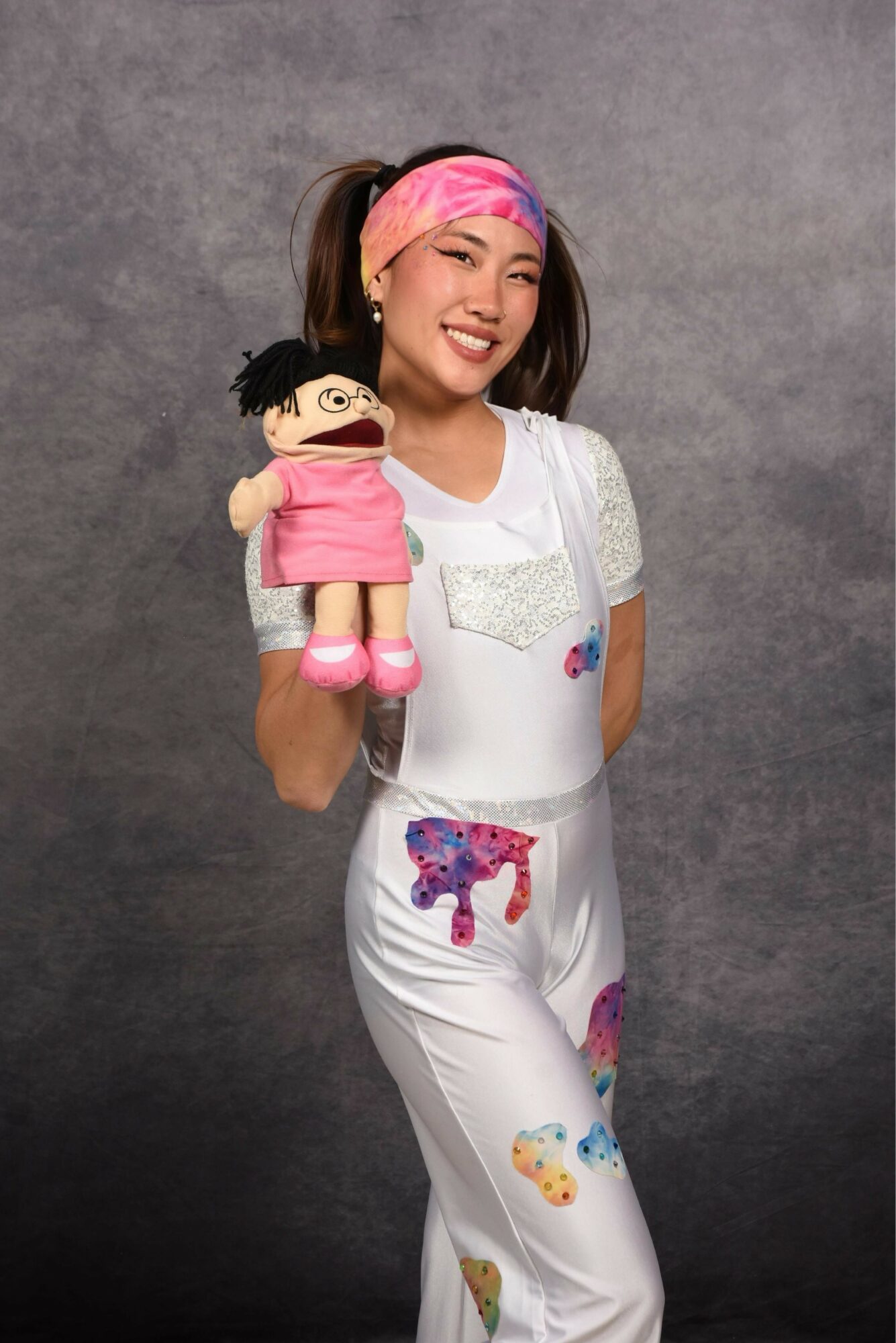 Young woman with a pink headband holding a puppet, smiling, standing against a gray background.