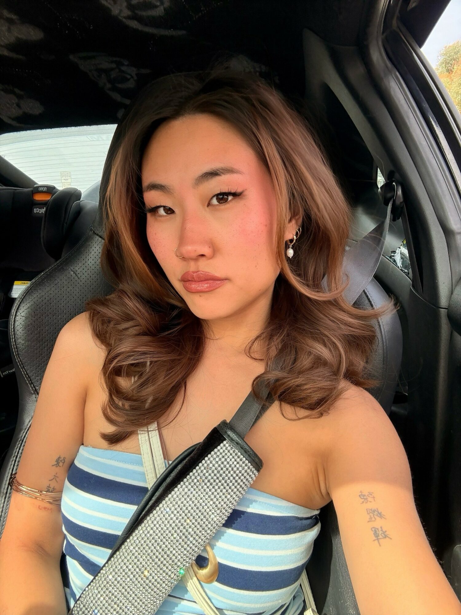Young woman with wavy hair sitting in a car, wearing a striped top and a seatbelt.
