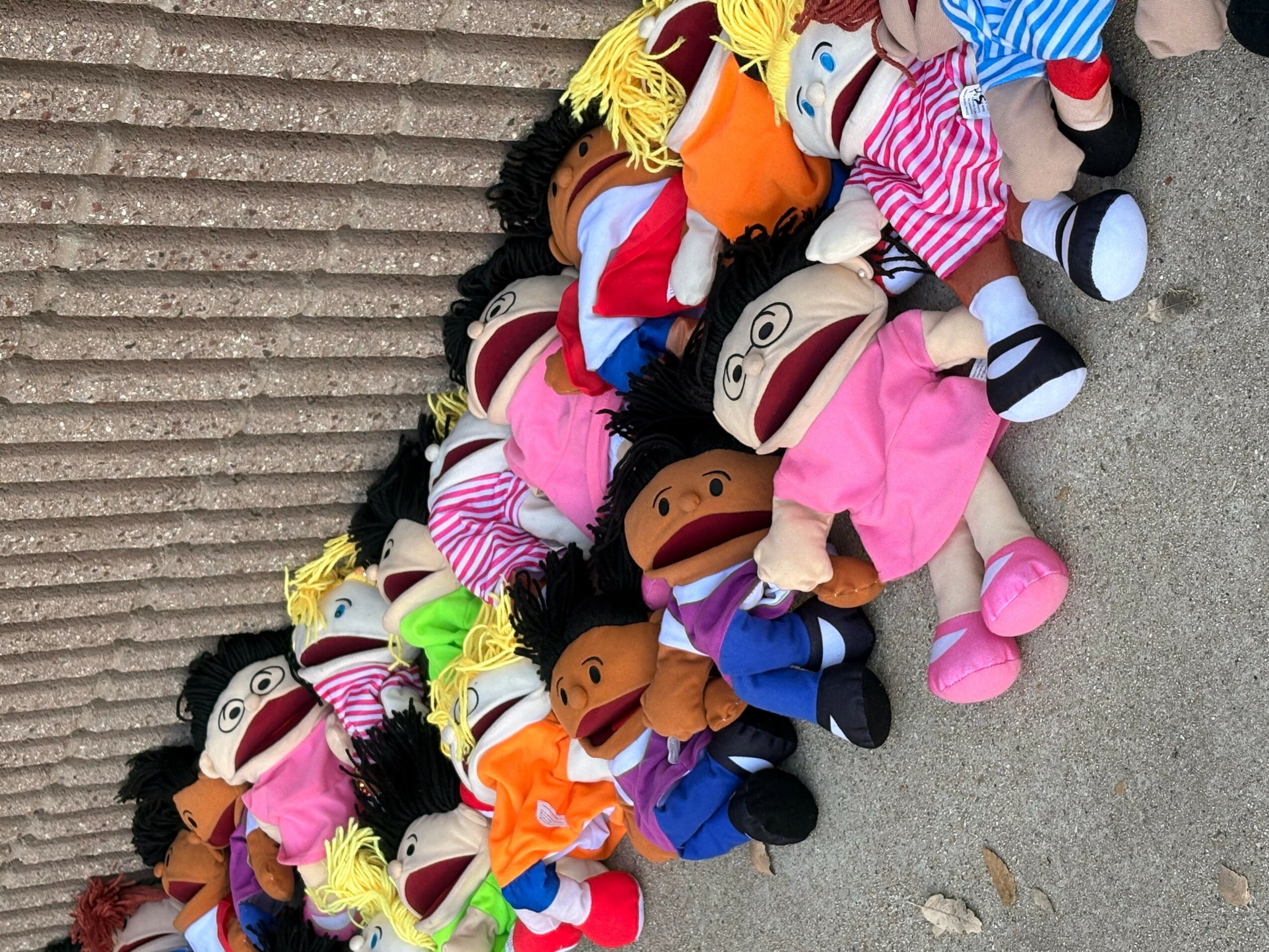 Pile of colorful plush dolls with various costumes and hairstyles on a concrete surface.