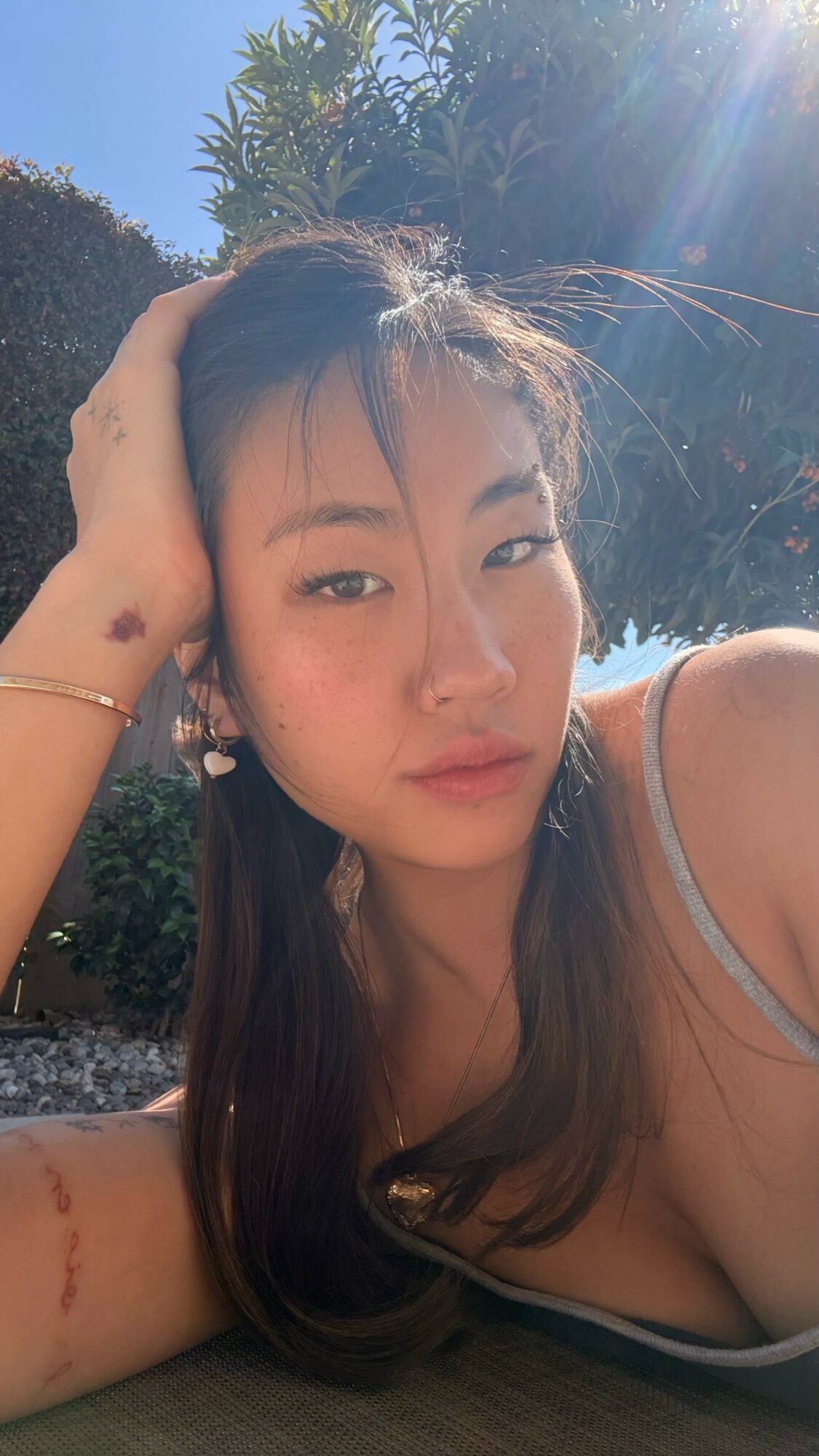 Young woman with long hair outdoors, sunlight on face, hand resting on head, wearing a tank top.