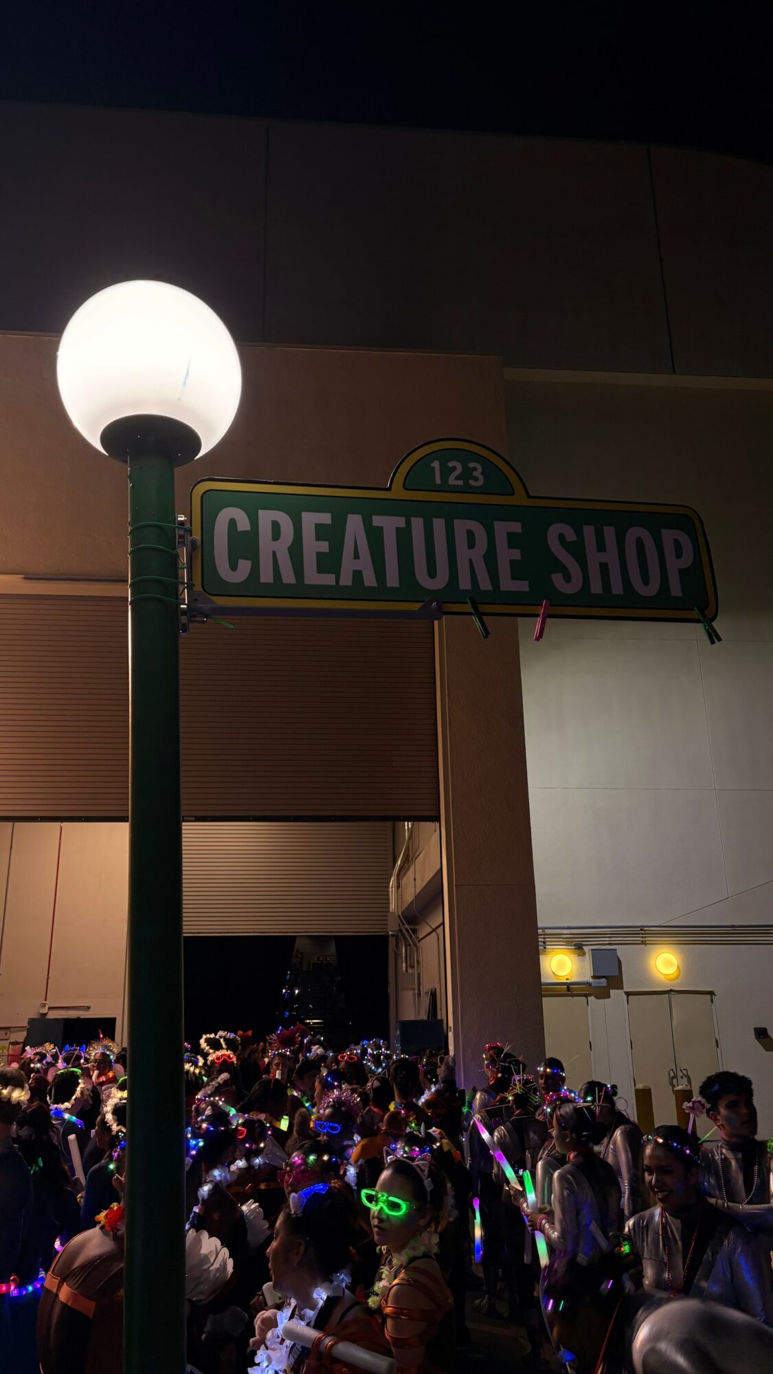 Street sign reading 'Creature Shop' illuminated by a nearby streetlamp at night, with a crowd of people below.
