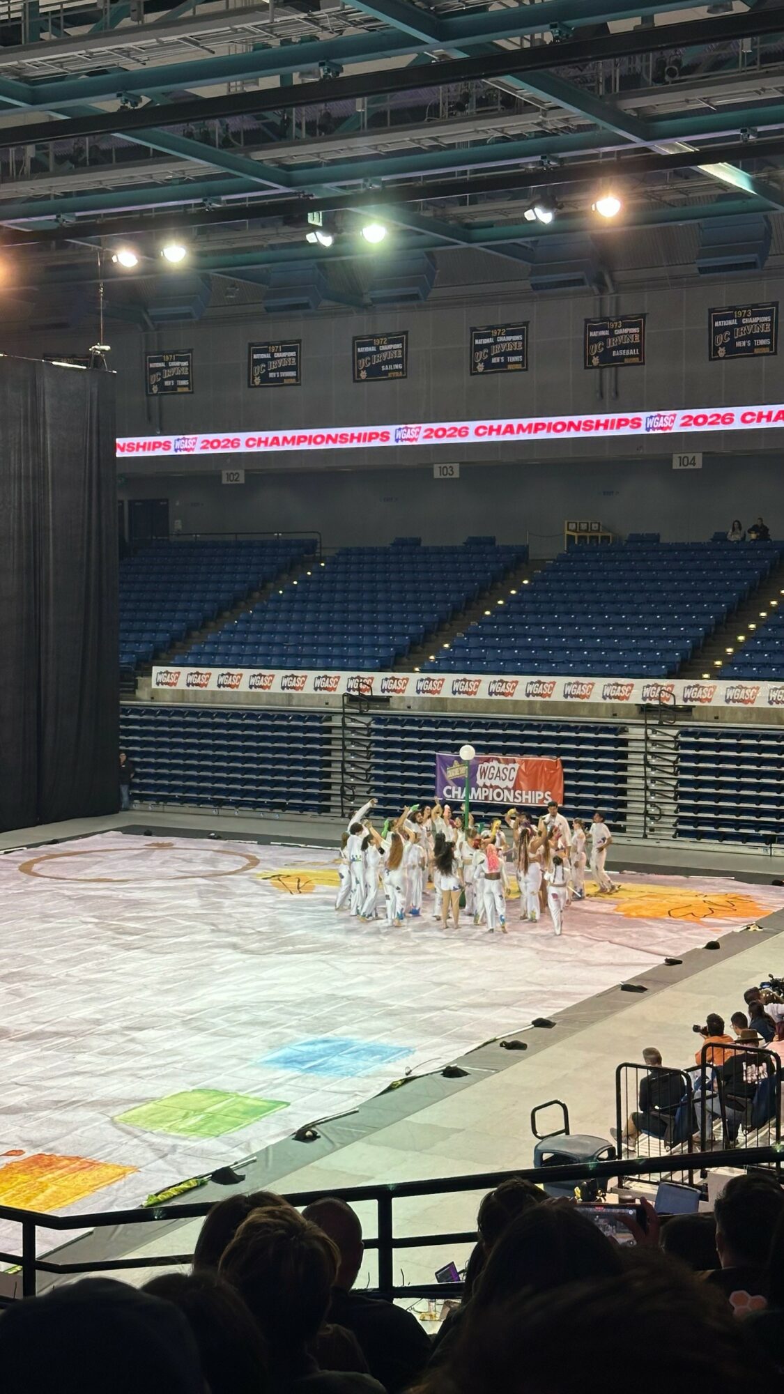 Group of figure skaters on ice rink in empty indoor arena with blue seats and overhead lights.