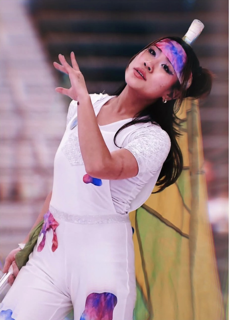 Young woman with colorful face paint and headband, wearing white outfit, posing with hand raised, indoors with colorful background.