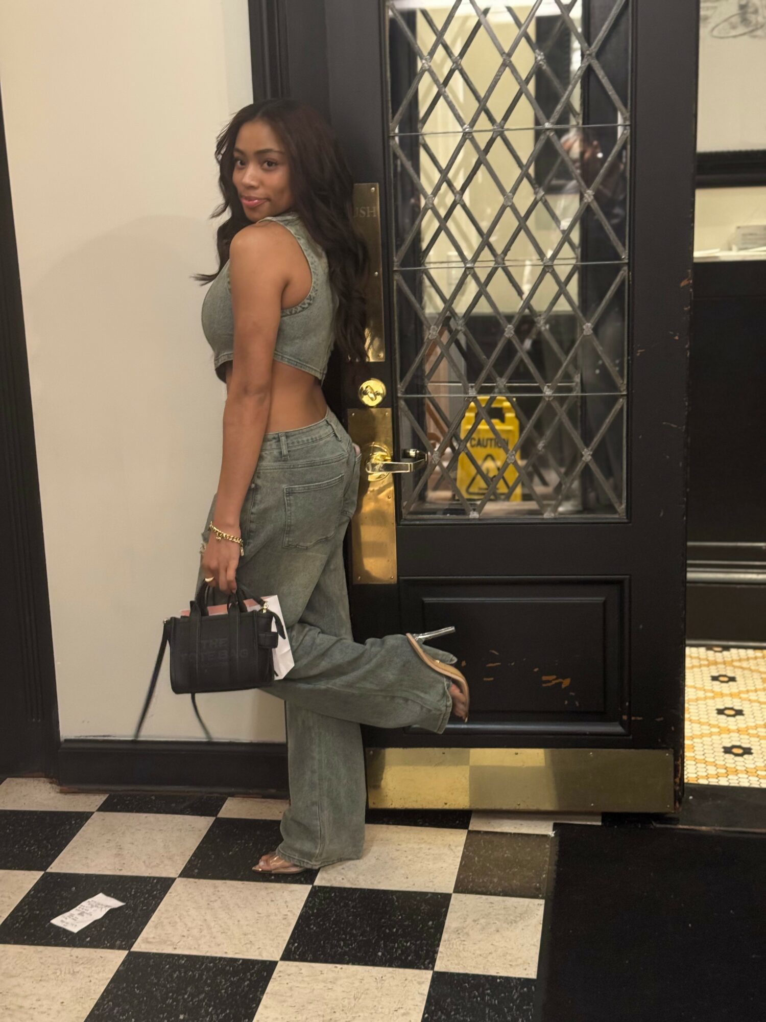 Woman standing in front of a glass door, holding a black handbag, wearing a gray crop top and jeans, smiling.
