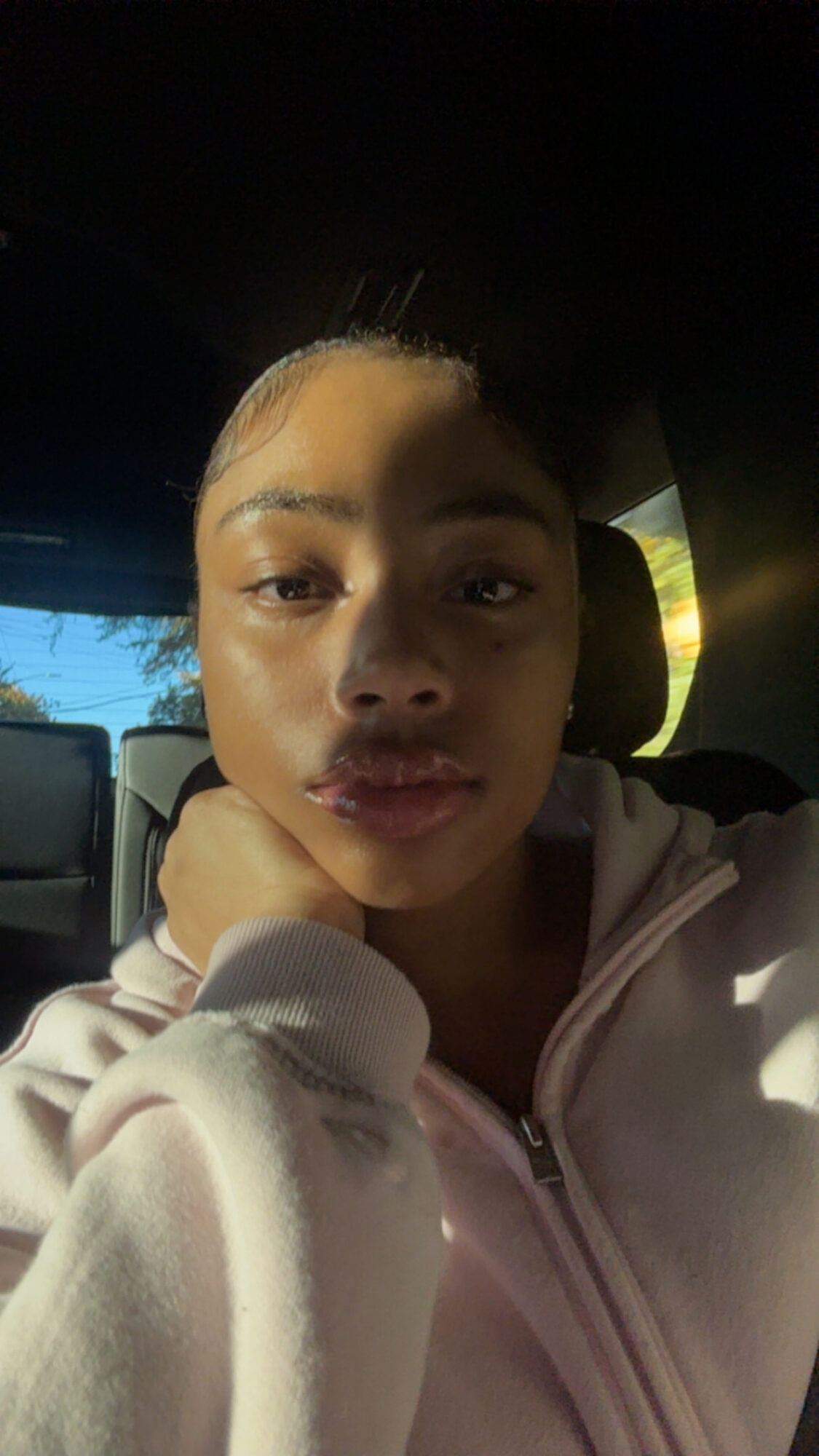 Young woman with light skin and blonde hair, sitting in a vehicle, resting her chin on her hand, looking at the camera.
