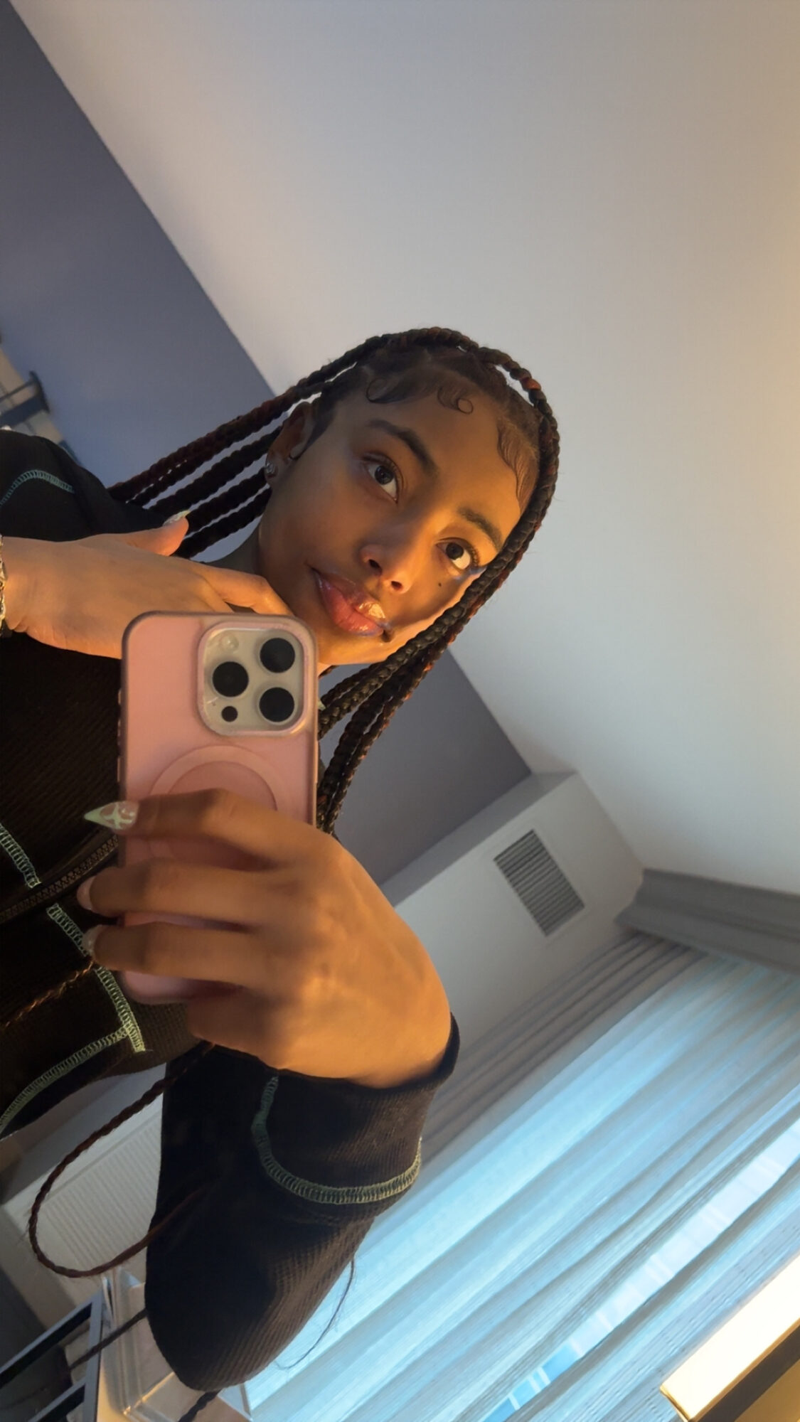 Young woman with braided hair taking a mirror selfie with a pink phone in a room with curtains.