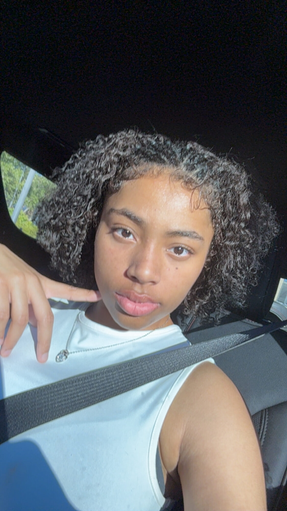 Young woman with curly hair taking a selfie in a car, wearing a white sleeveless top, seatbelt visible.