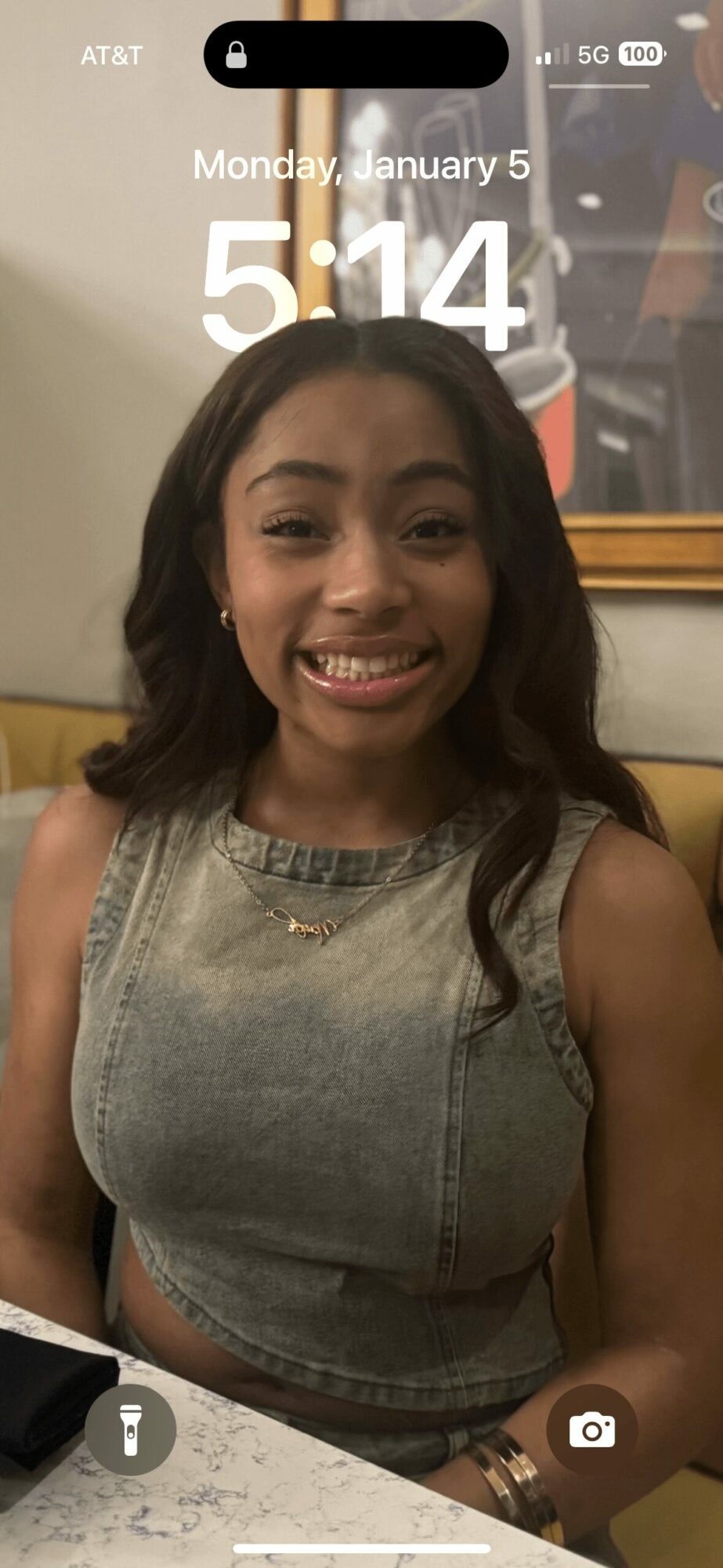 Young woman with dark hair smiling, wearing a sleeveless gray top, sitting indoors at a table.