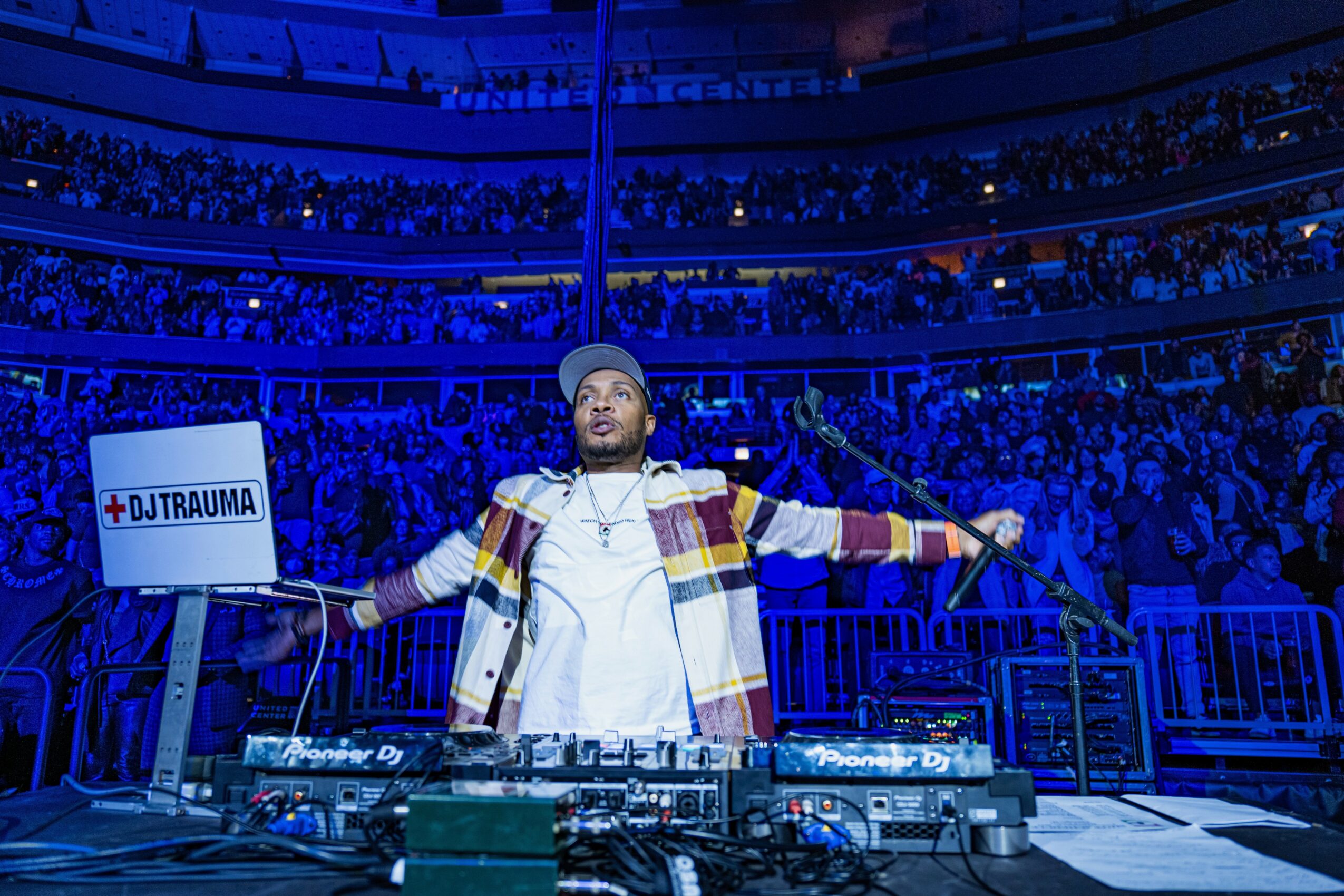DJ performing on stage with arms outstretched in front of a large audience in a stadium.