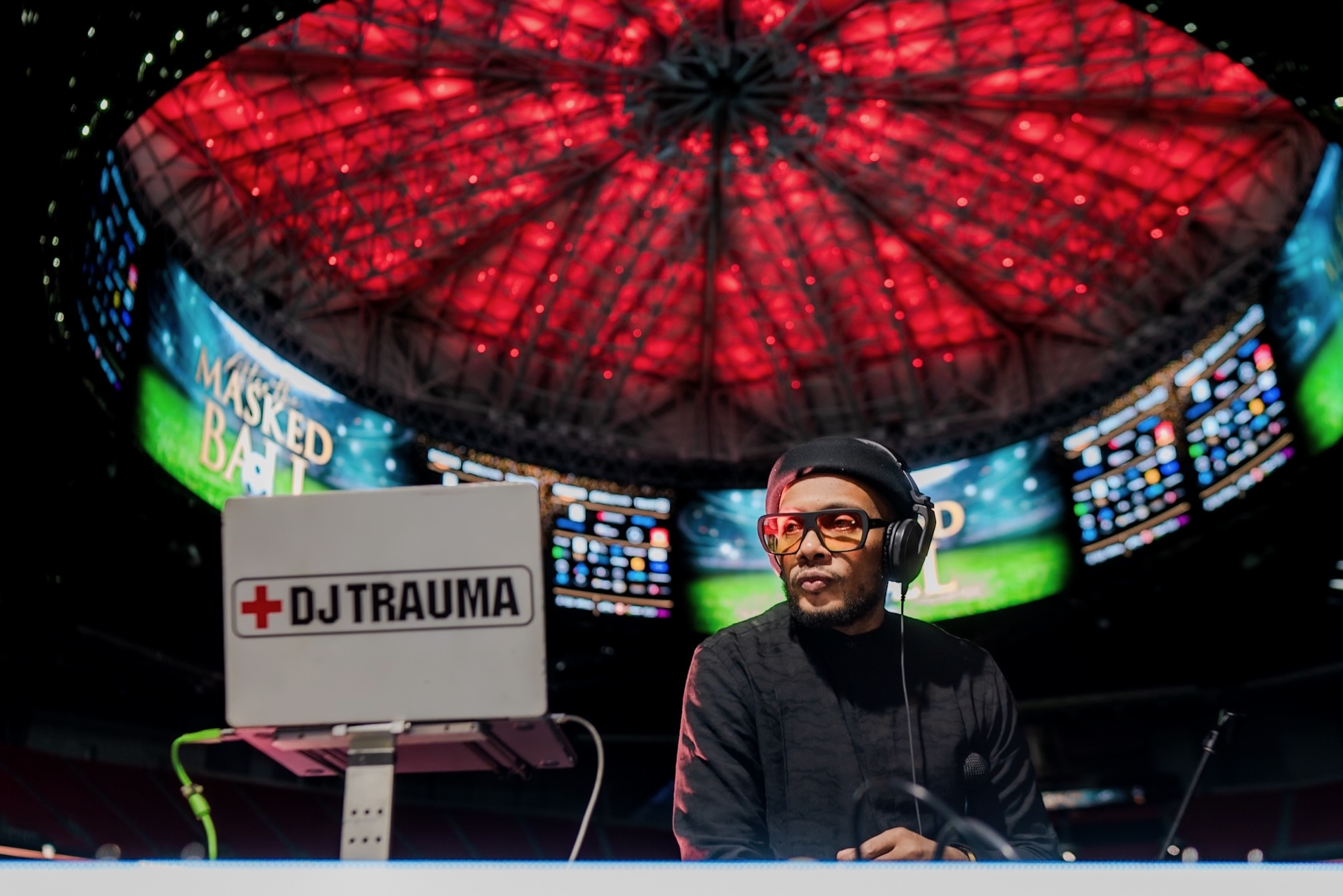 DJ wearing headphones and glasses, smiling, in front of a screen with a red circular ceiling structure above.