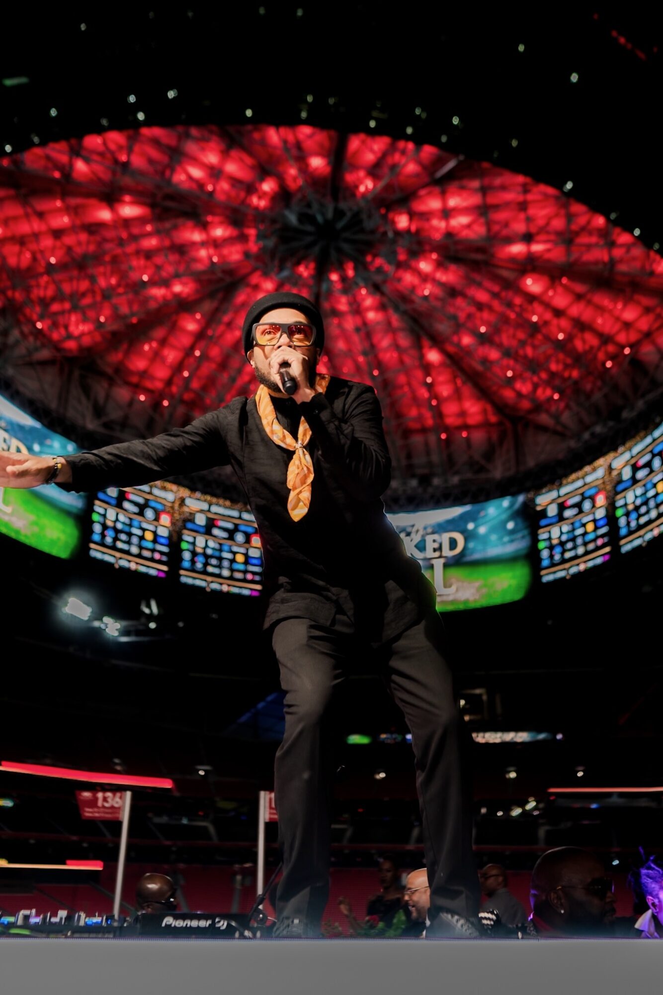 Person performing on stage with a large red-lit circular ceiling above, surrounded by screens displaying colorful graphics.