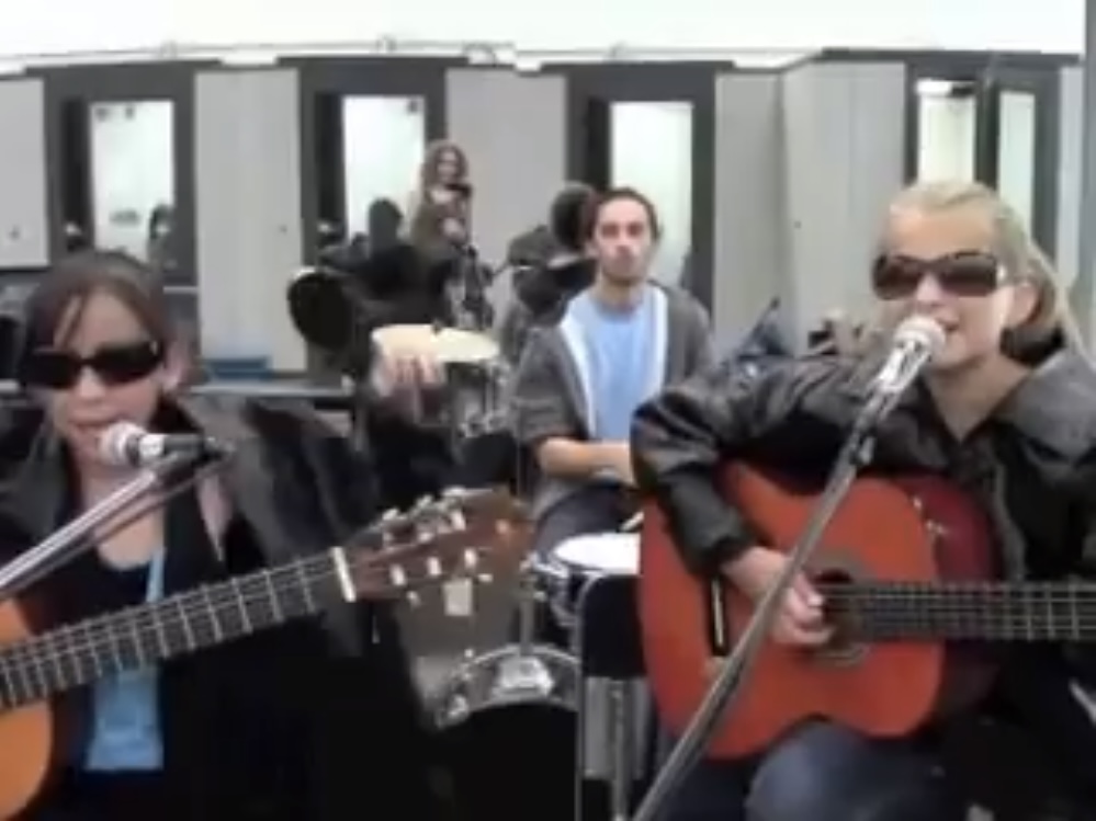Group of people playing guitars and singing outdoors, with a mirror wall behind them.