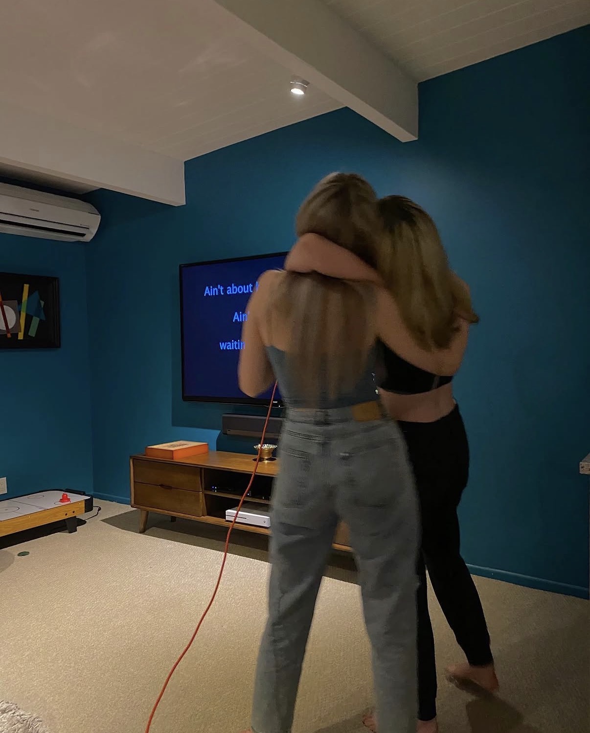 Two women hugging in a room with a blue wall and a television screen.