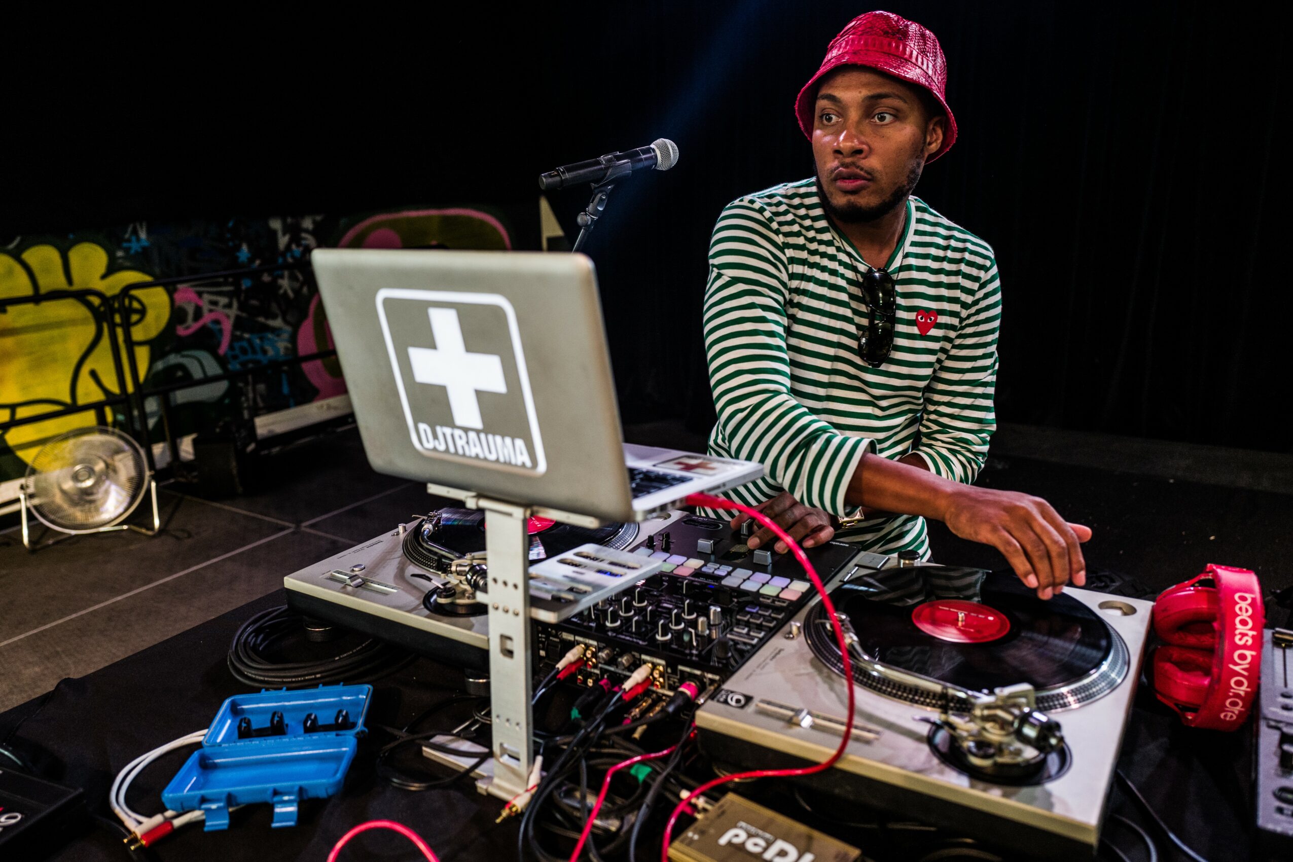 Person wearing striped shirt and red hat operating DJ equipment on stage with microphone nearby.