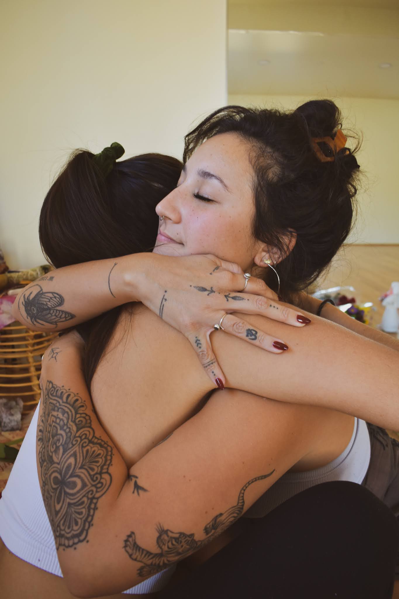 Two women hugging closely, one with tattoos on arms and shoulder, the other with curly hair, in an indoor setting.