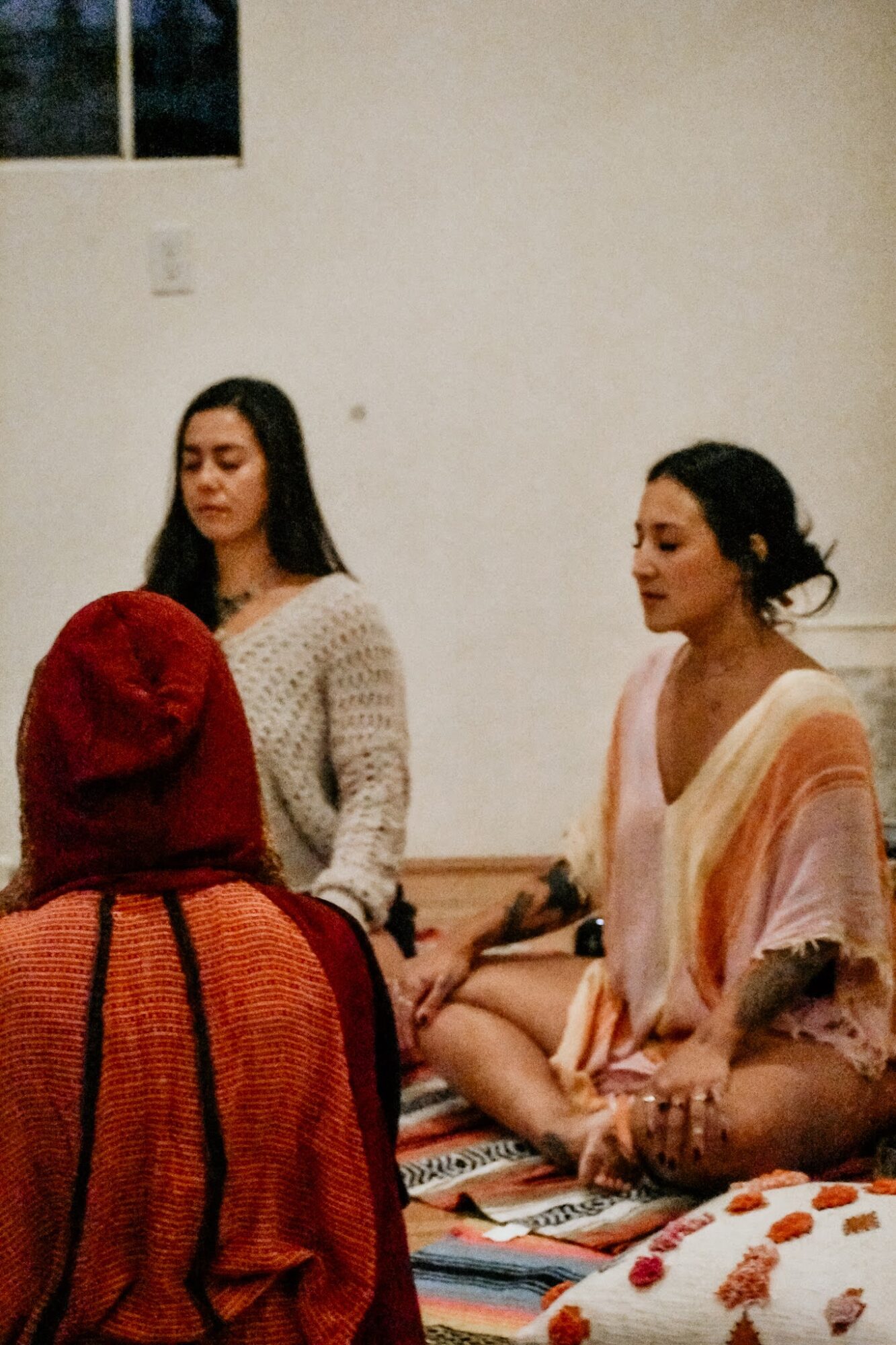 Two women sit on a colorful striped mat, one with dark hair in a bun and the other with long dark hair, in a room with plain white walls. A person with a red head covering is in the foreground.