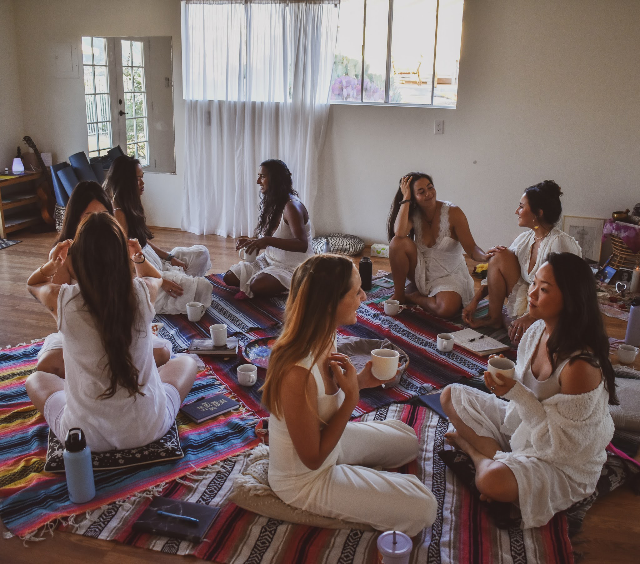 Group of women sitting on colorful rugs in a bright room, engaging in conversation and activities.