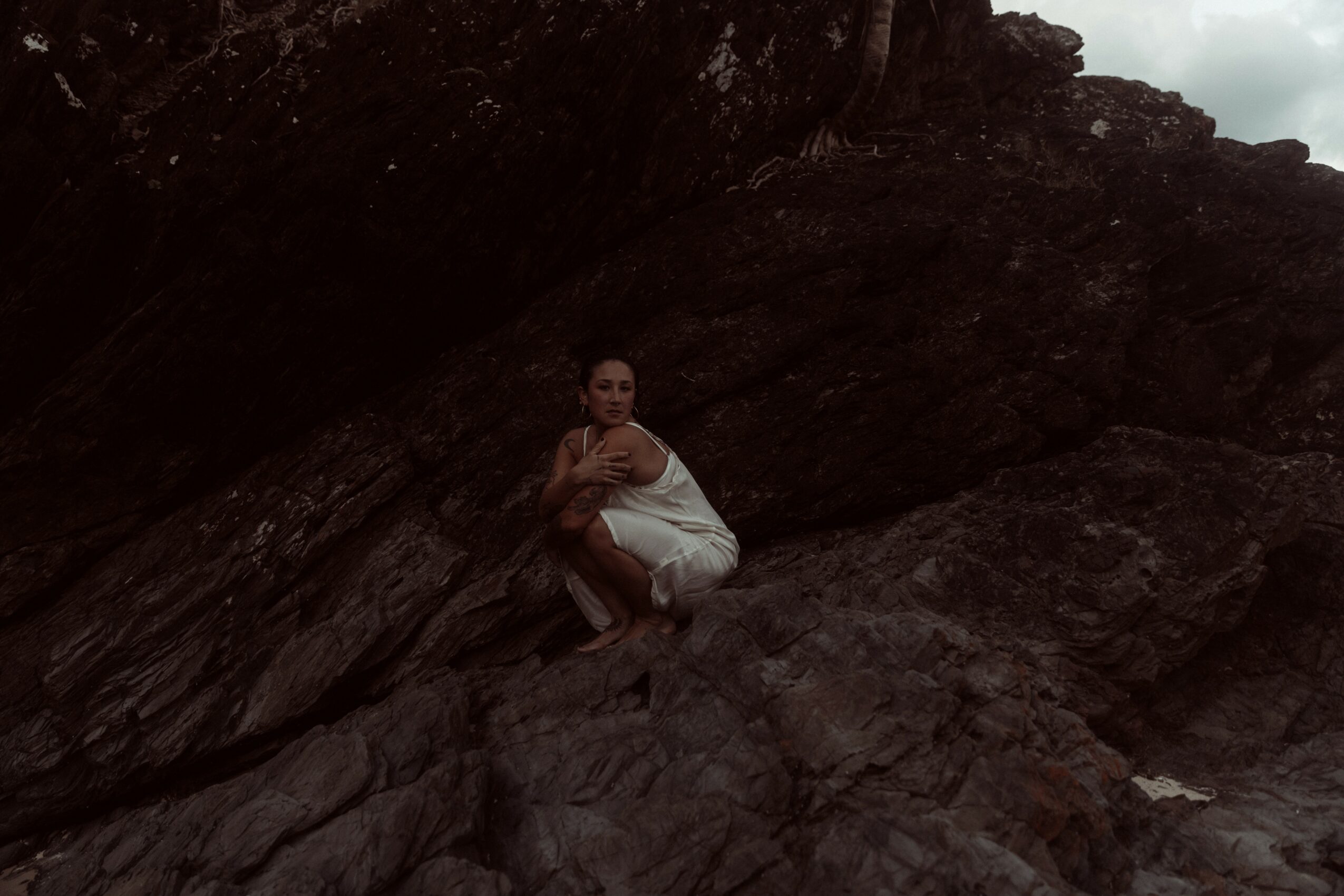 Person sitting on dark rocks outdoors, wearing white clothing, with a rocky background and cloudy sky.