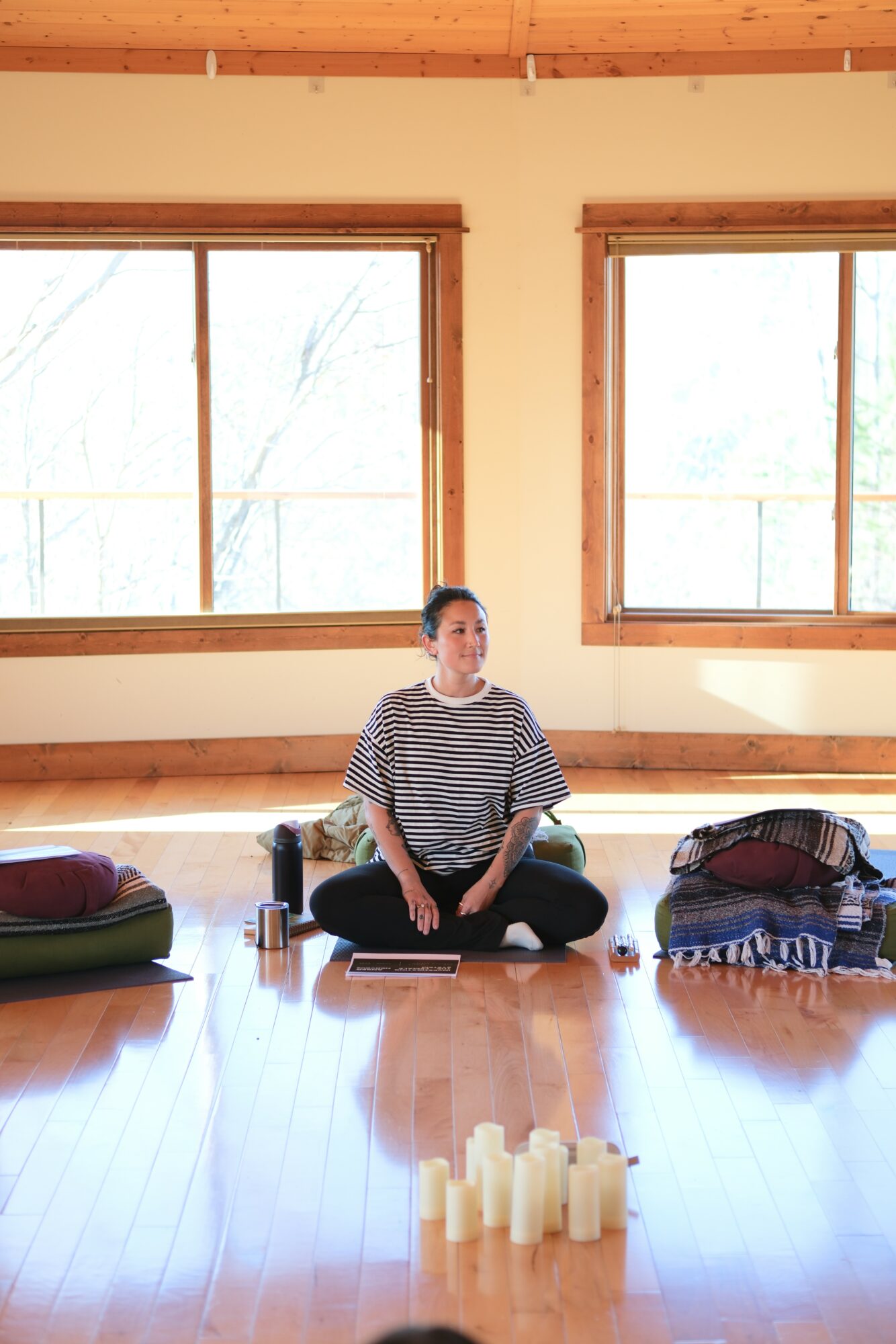 Person sitting cross-legged on a mat in a room with wooden floors and large windows, surrounded by bags and candles.