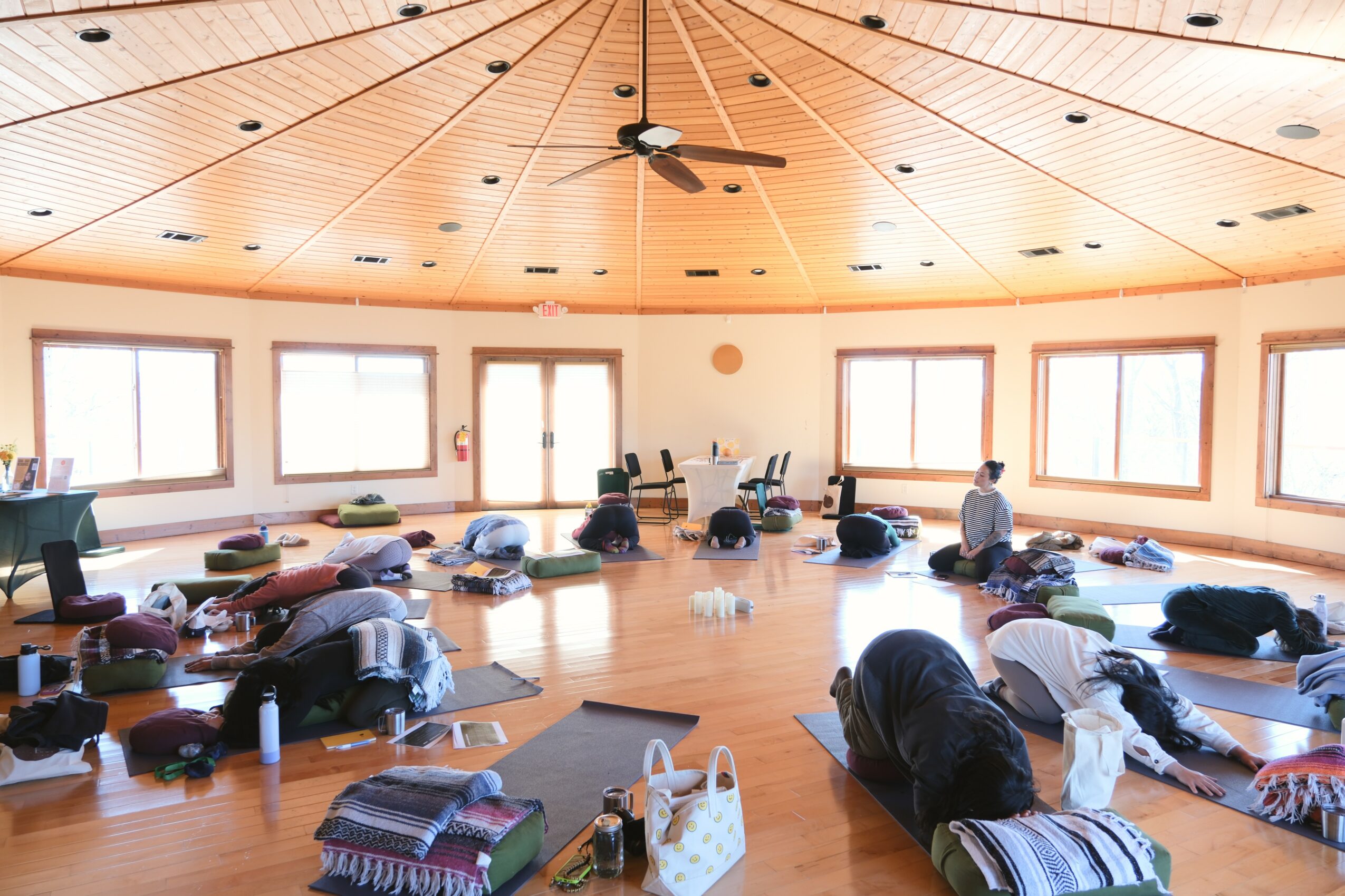 People practicing yoga or meditation in a spacious, well-lit room with large windows and wooden ceiling.