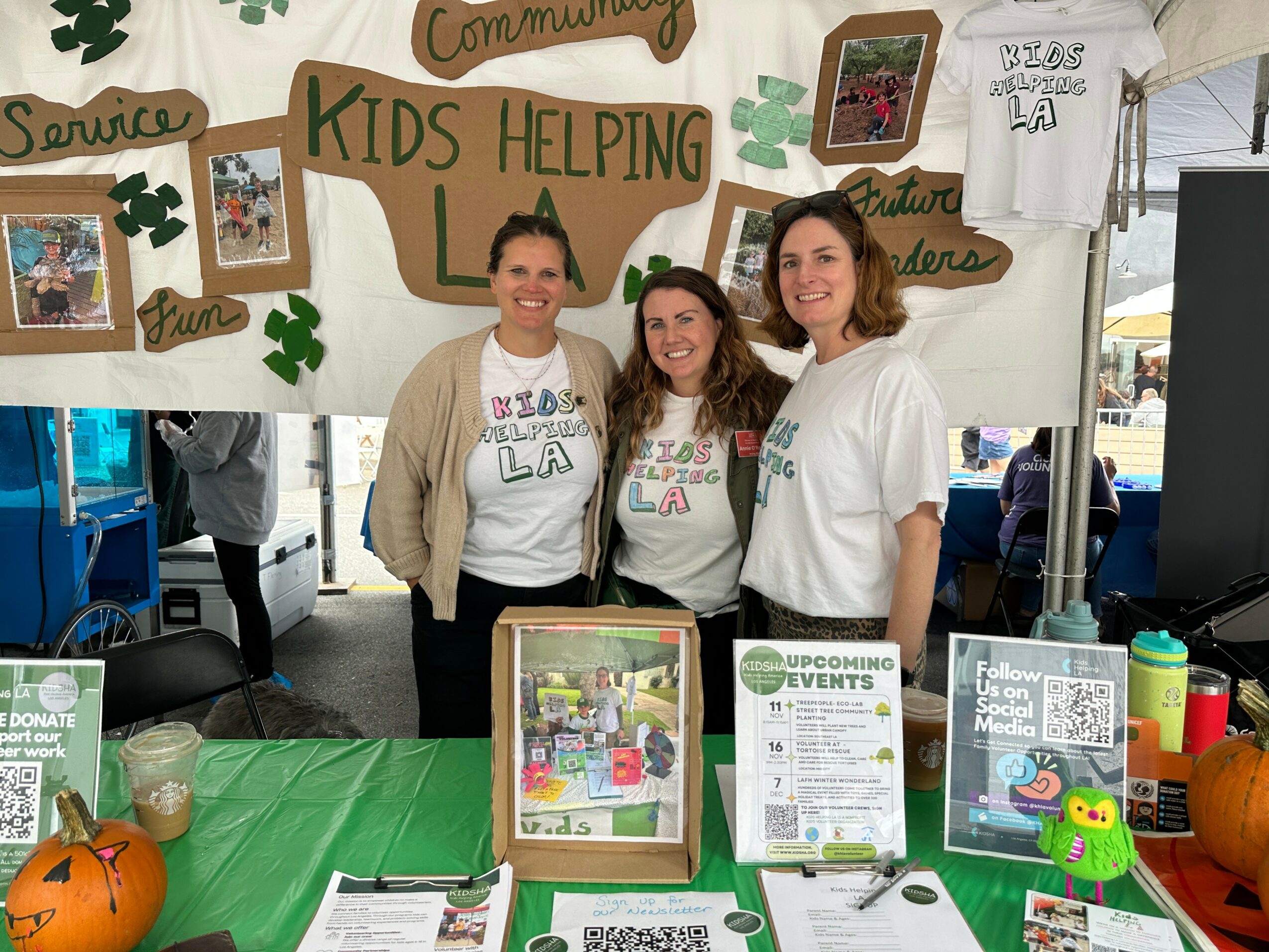 Three women standing behind a table at a community event, smiling, with a banner reading 'Kids Helping Kids' and various signs.
