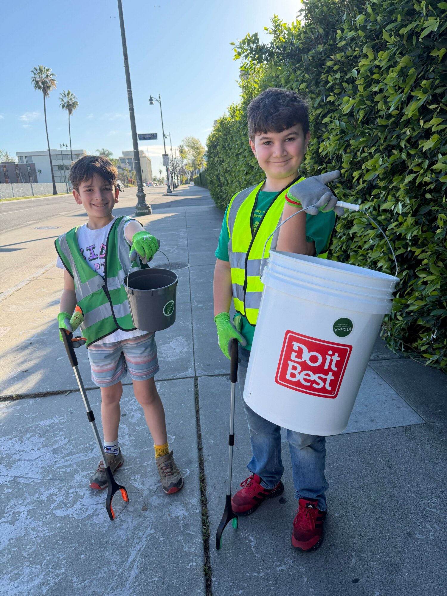 Two boys in green safety vests holding trash pickers and buckets, smiling outdoors on a sunny day.