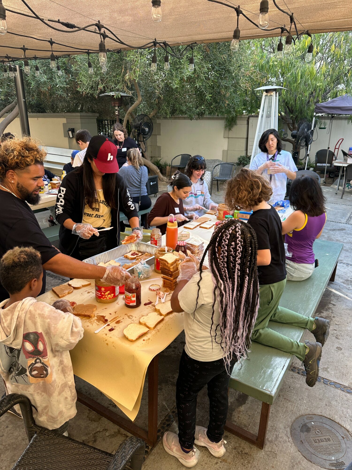 Group of people gathered around a table outdoors, eating and socializing under a canopy with trees in background.