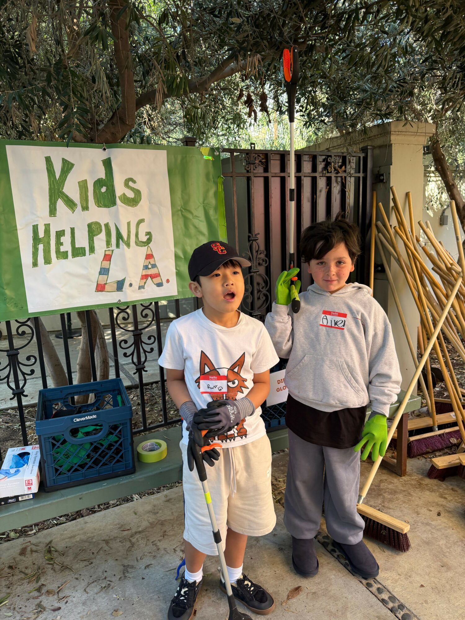 Two children standing outdoors near a fence, holding cleaning tools, with a green banner reading 'Kids Helping LA'.