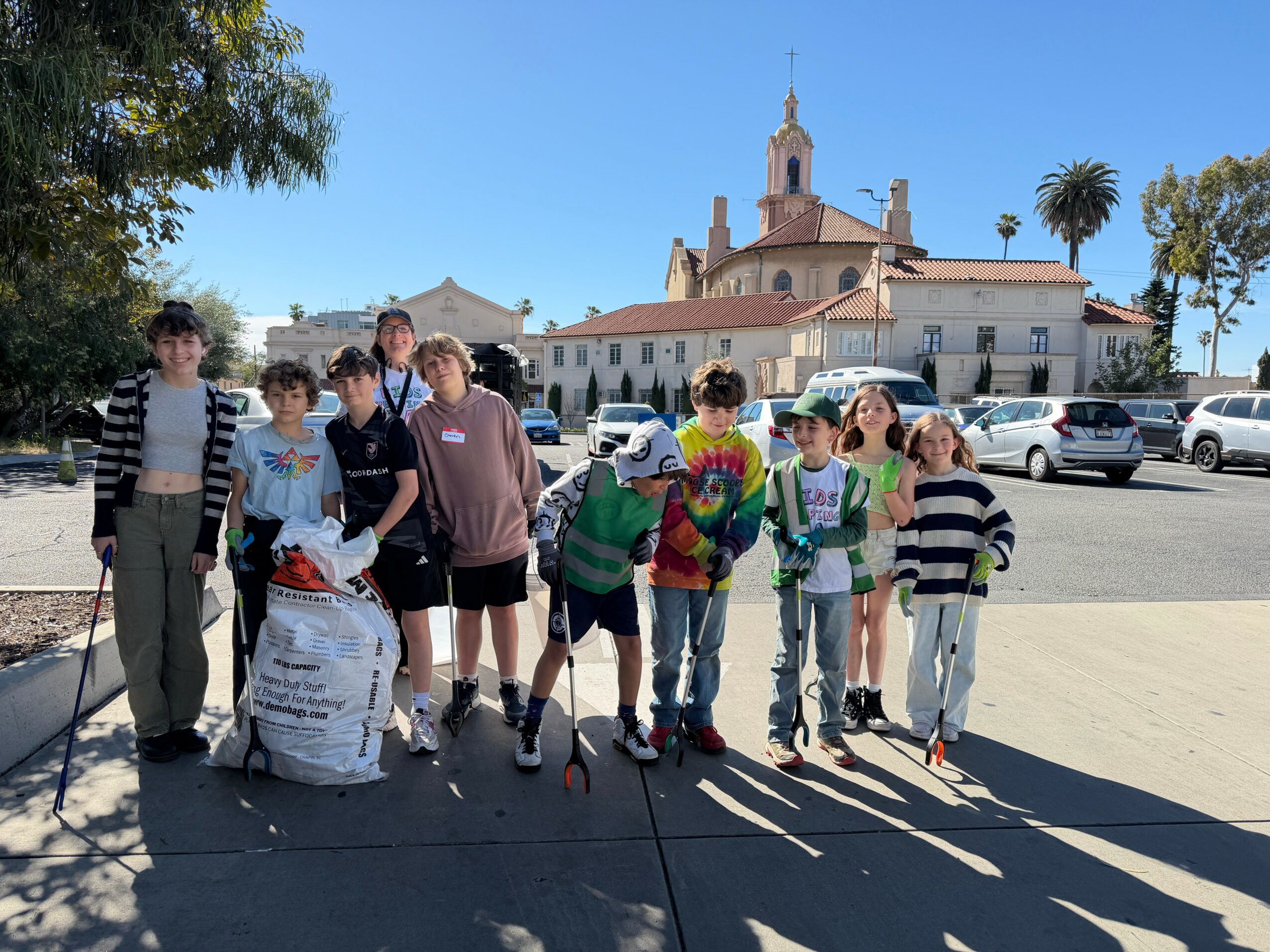 Group of children standing outdoors in front of a church, some holding bags and backpacks, with cars parked behind.