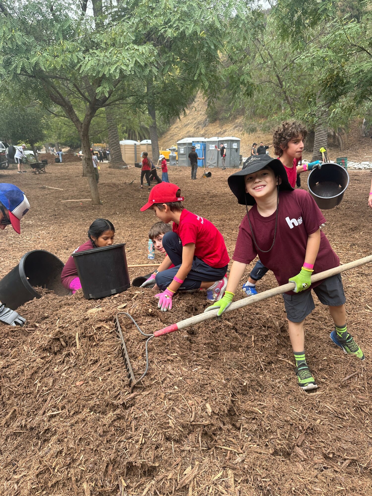 Children and adults planting trees outdoors with shovels and tools, surrounded by trees and a park setting.