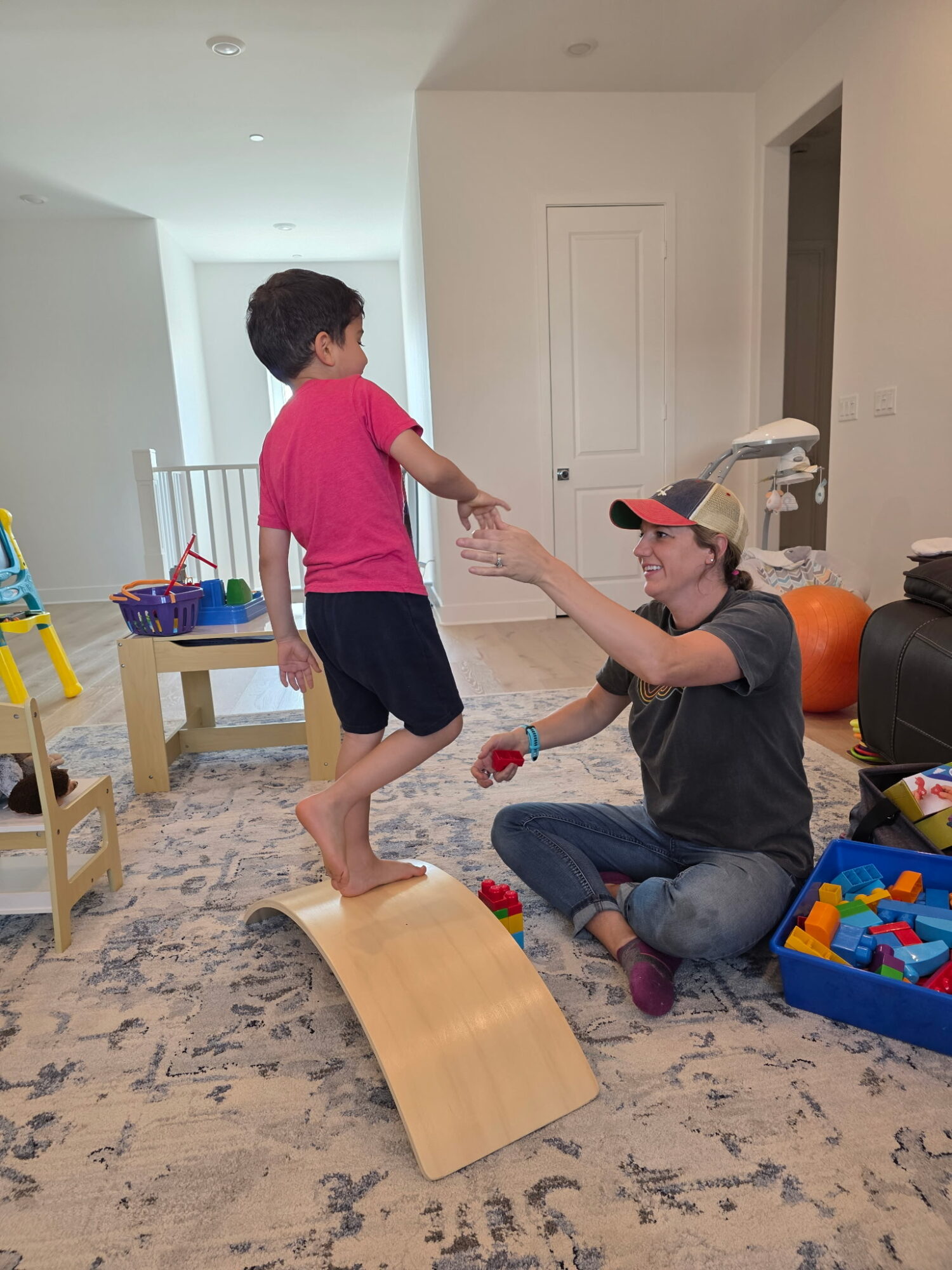 Child in pink shirt and shorts standing on a wooden ramp, reaching out to a woman sitting on the floor.