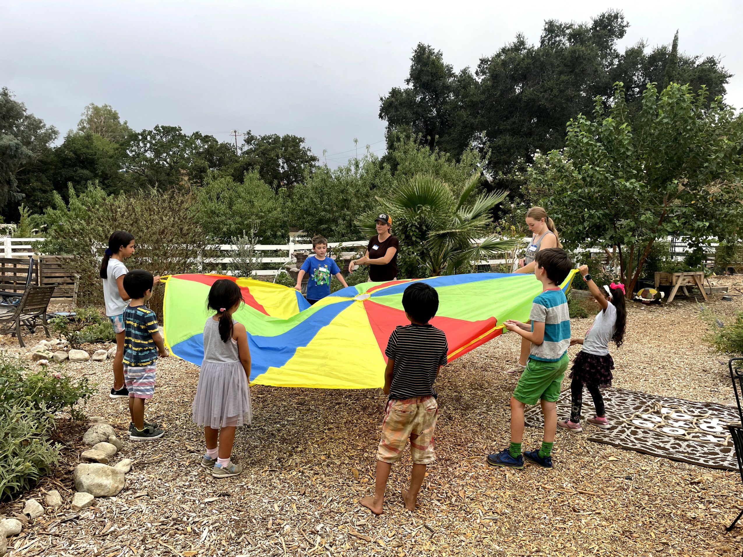 Children and an adult hold a colorful parachute outdoors in a garden area with trees and a white fence.