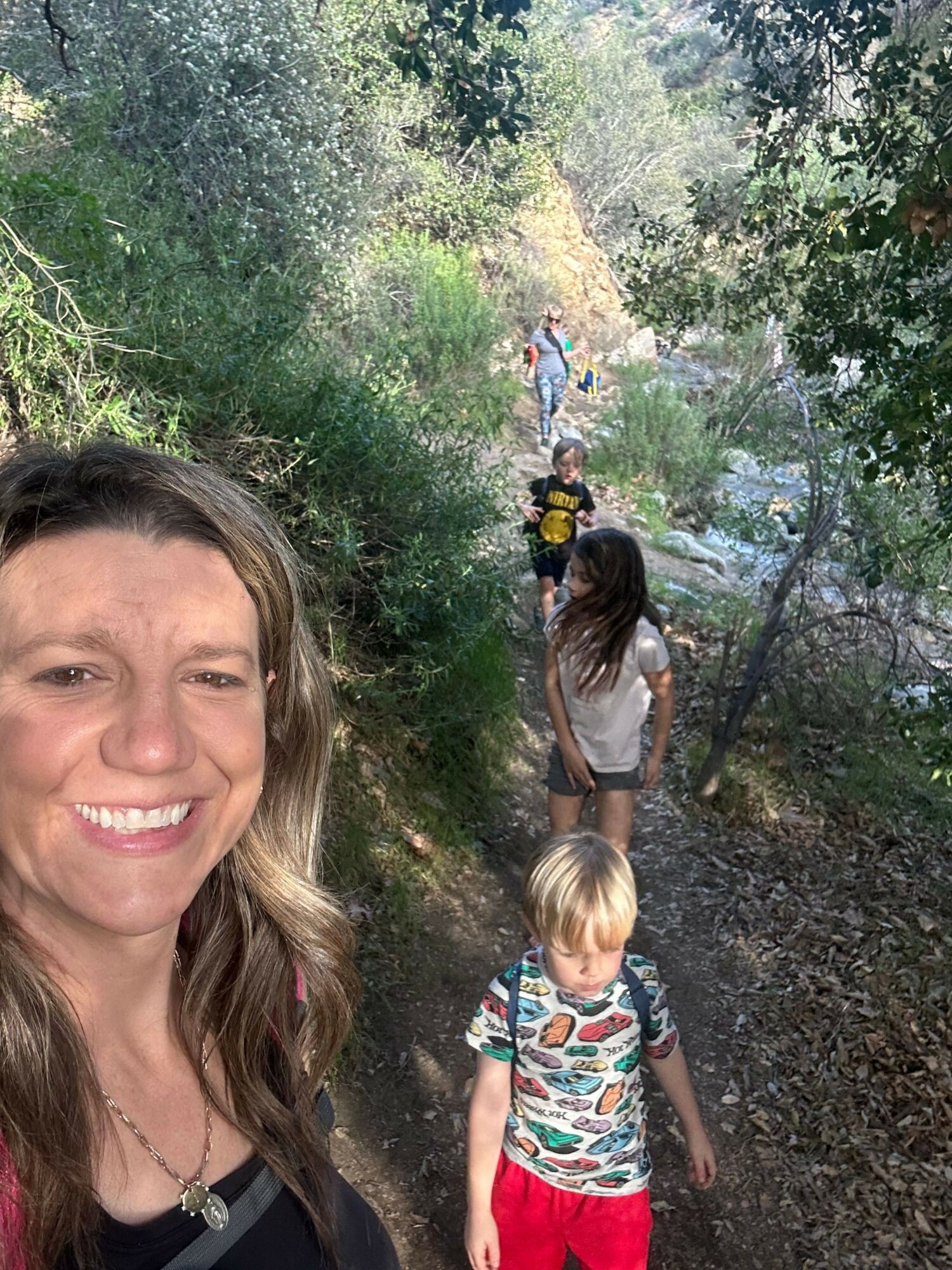 Group of people walking along a wooded trail outdoors, with trees and greenery around.