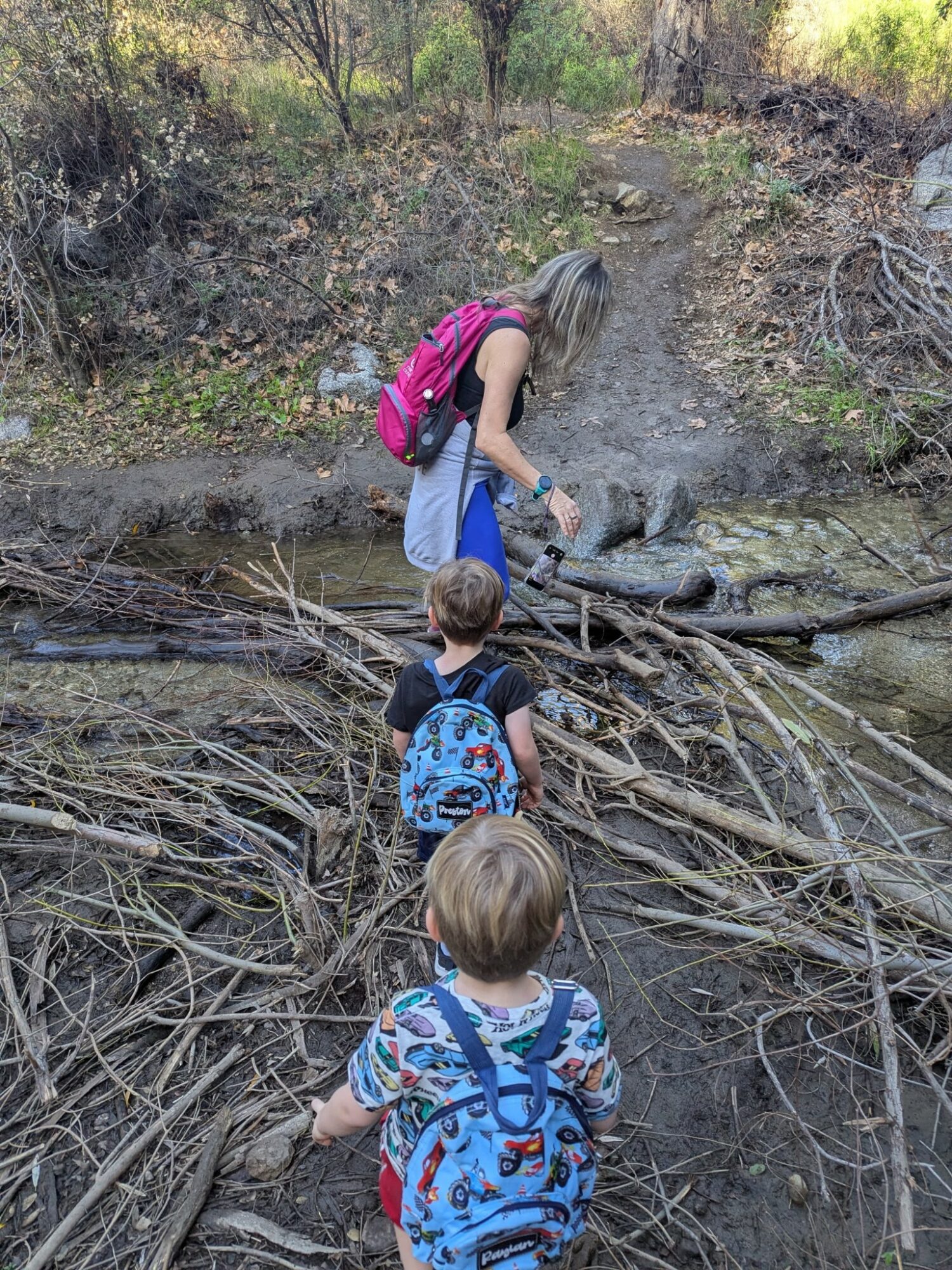 Two children with backpacks stand near a small stream in a wooded area, with a woman leaning over the water.