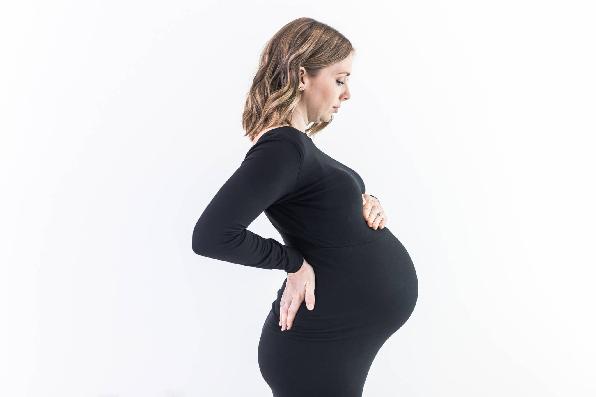 Pregnant woman in black dress standing sideways with hand on hip, profile view, white background.