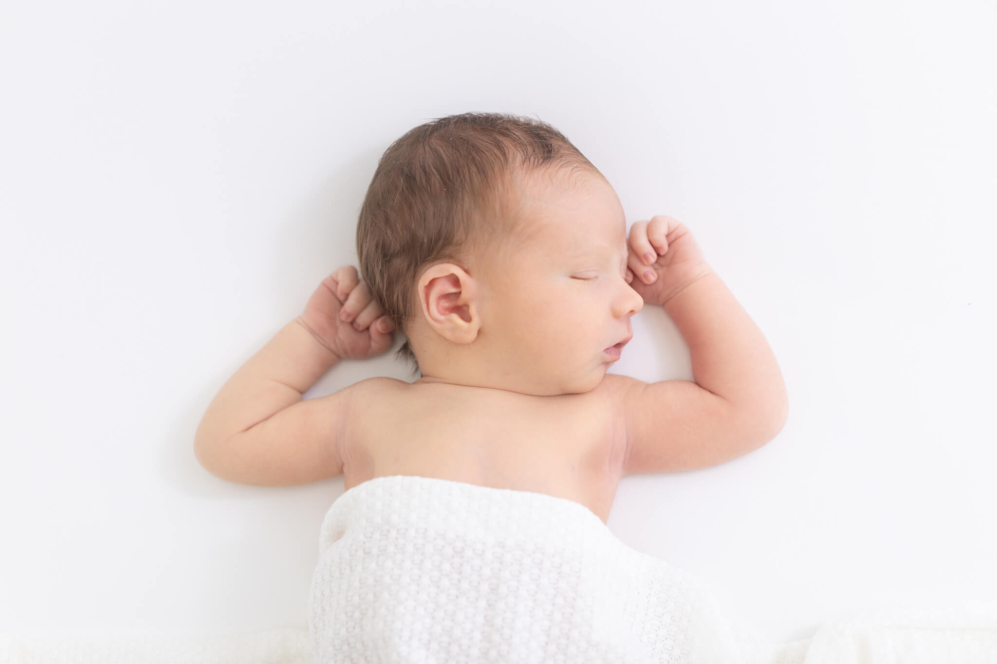 Baby sleeping on white surface, covered with a white blanket, with arms bent near head.