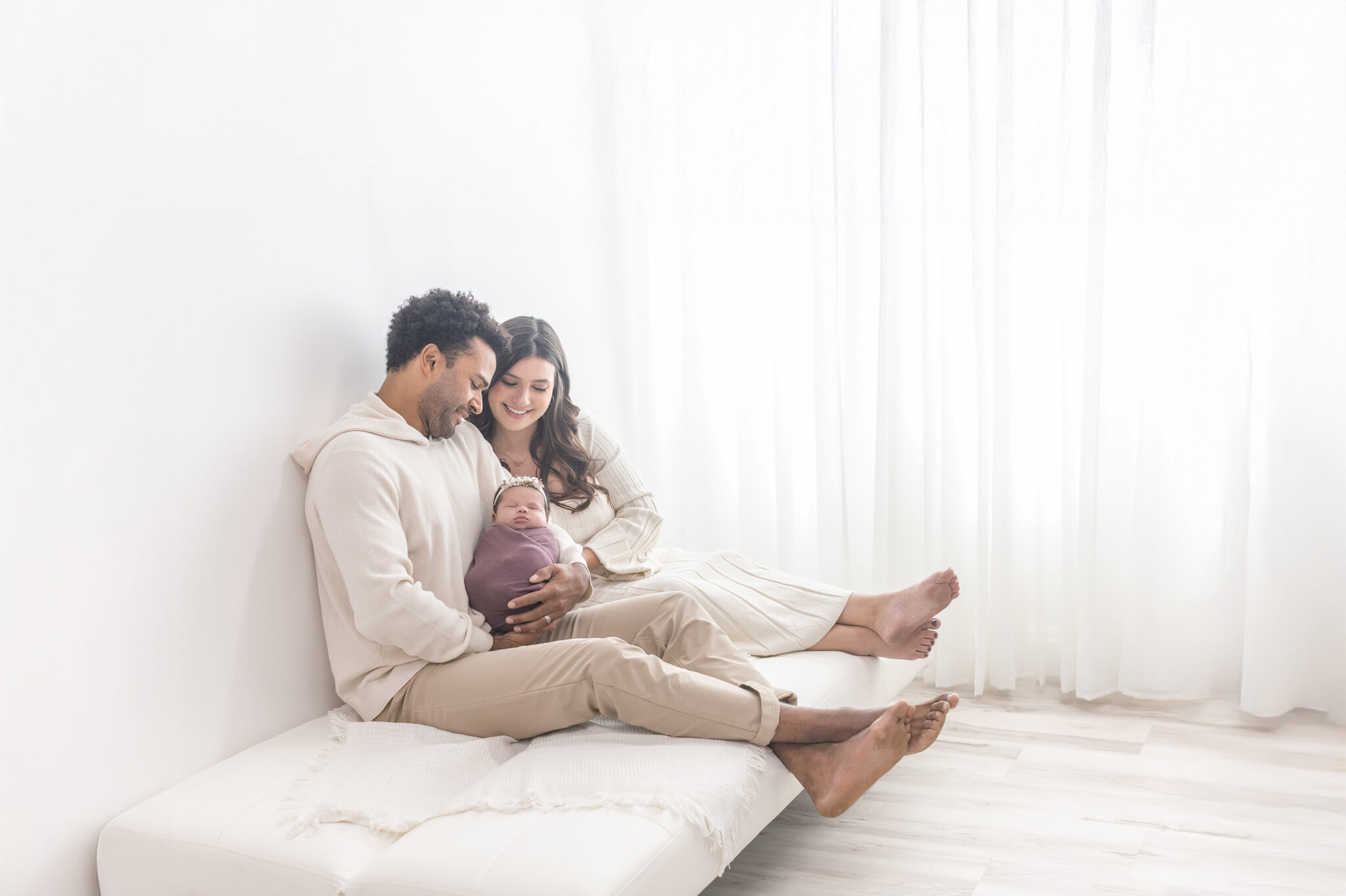 Family sitting on bed, with a pregnant woman and a man, smiling and looking at the woman’s belly, bright room.