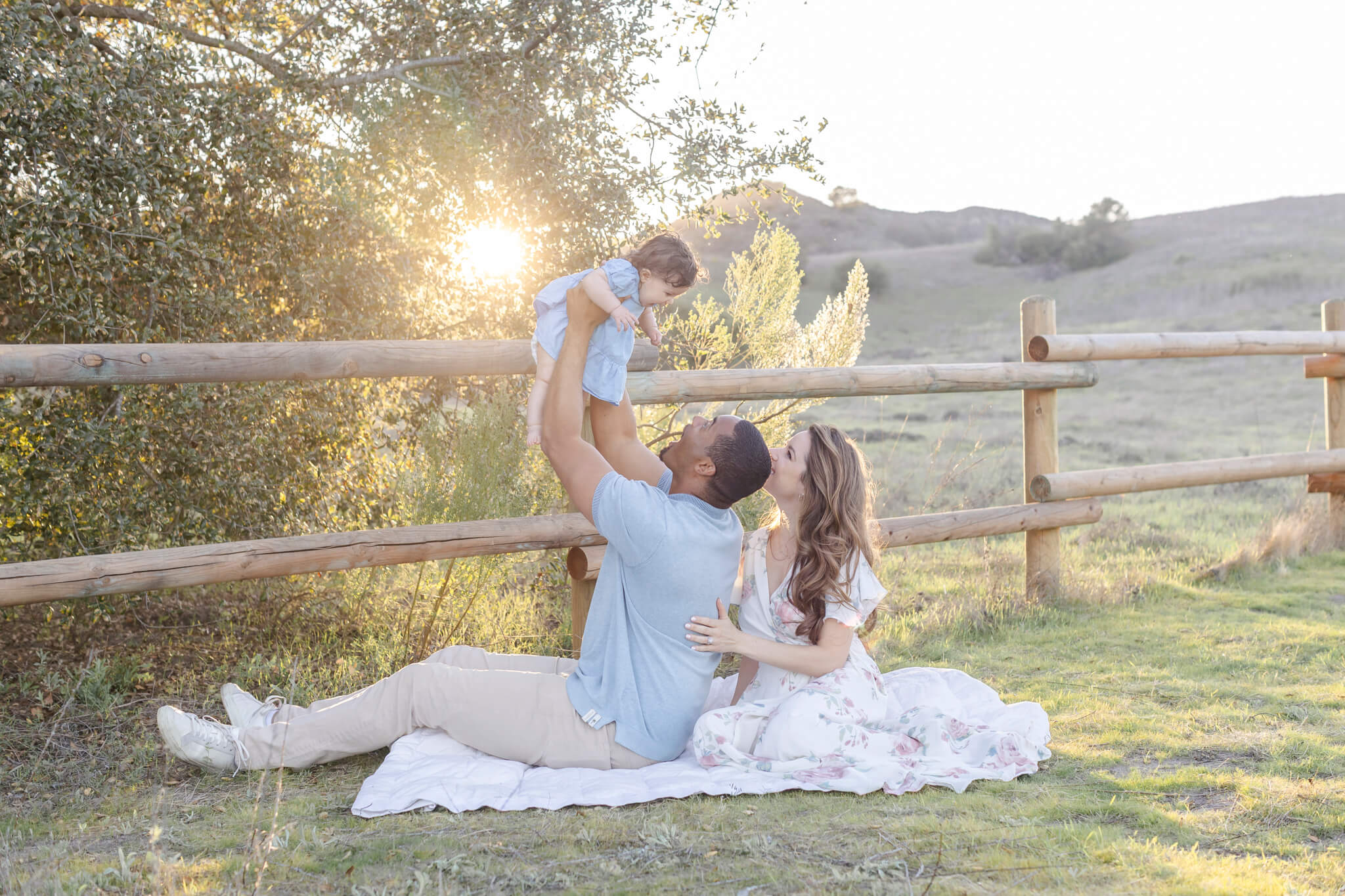 Man and woman sitting on a blanket outdoors, lifting a child in the air with a fence and trees in background.